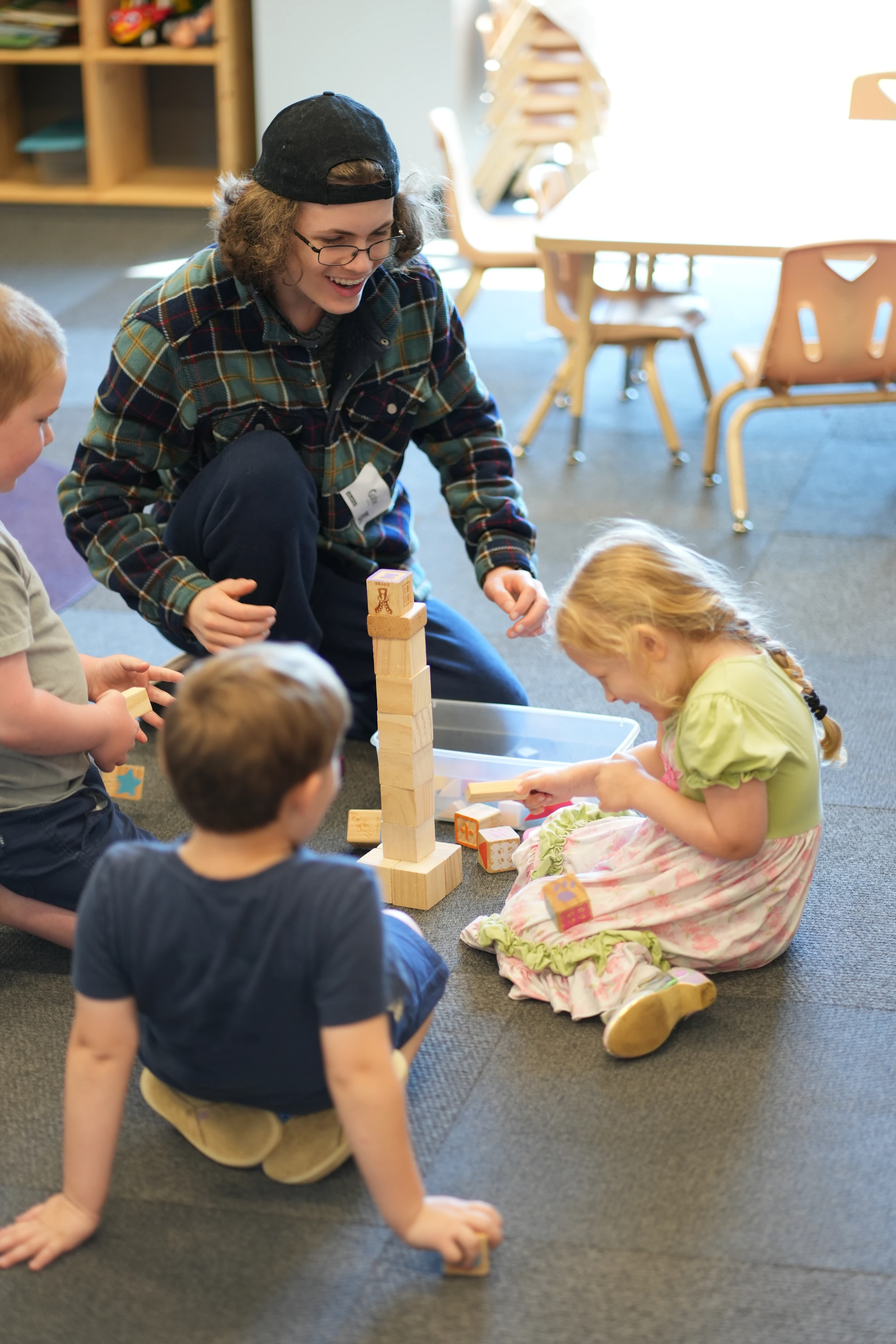 A smiling teacher sits on the floor with three young children, all playing together with wooden building blocks and making a tall tower in a classroom.