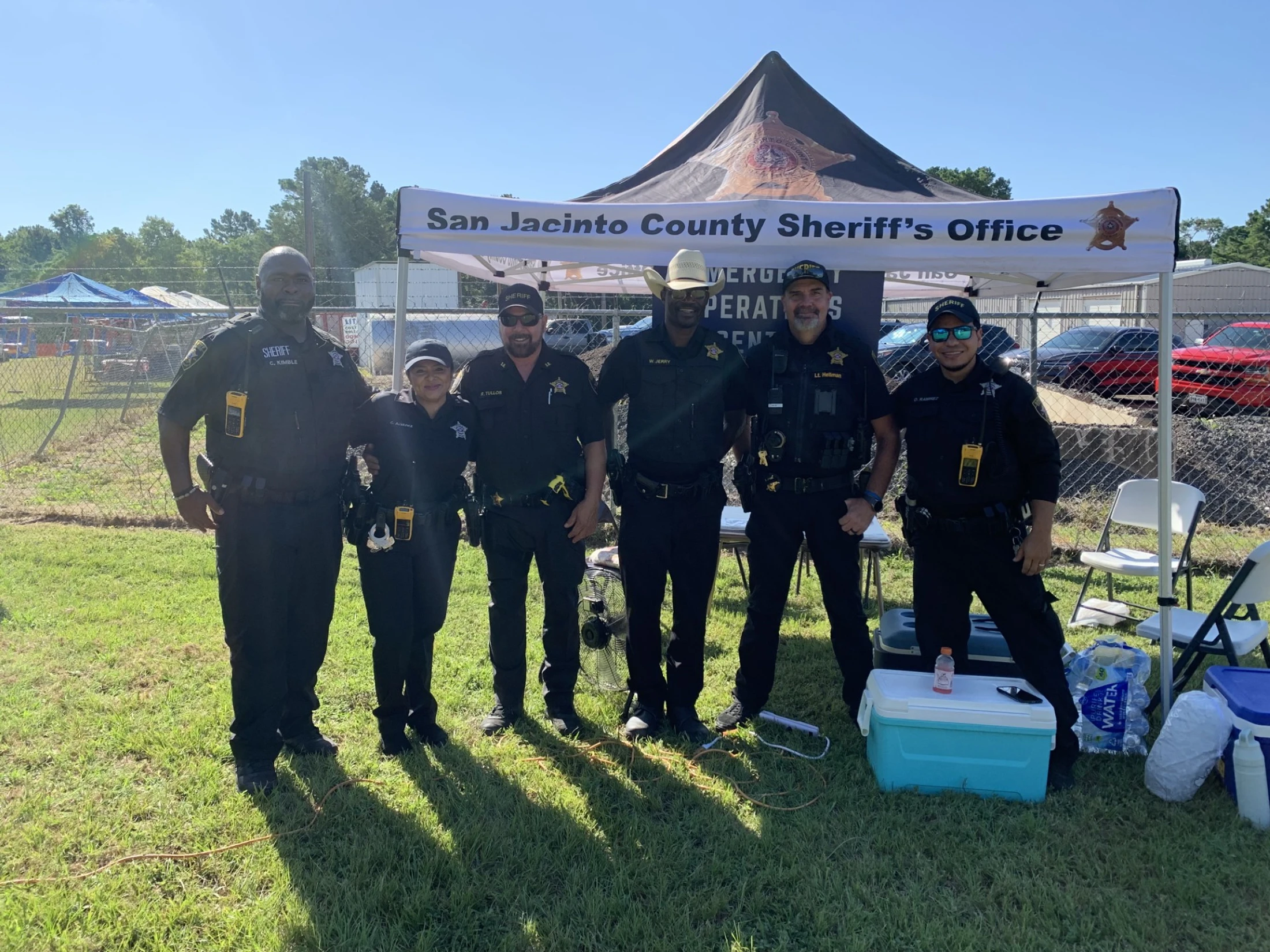 Six San Jacinto County Sheriff's deputies smile for a group photo in uniform, standing on grass in front of a branded tent on a sunny day.