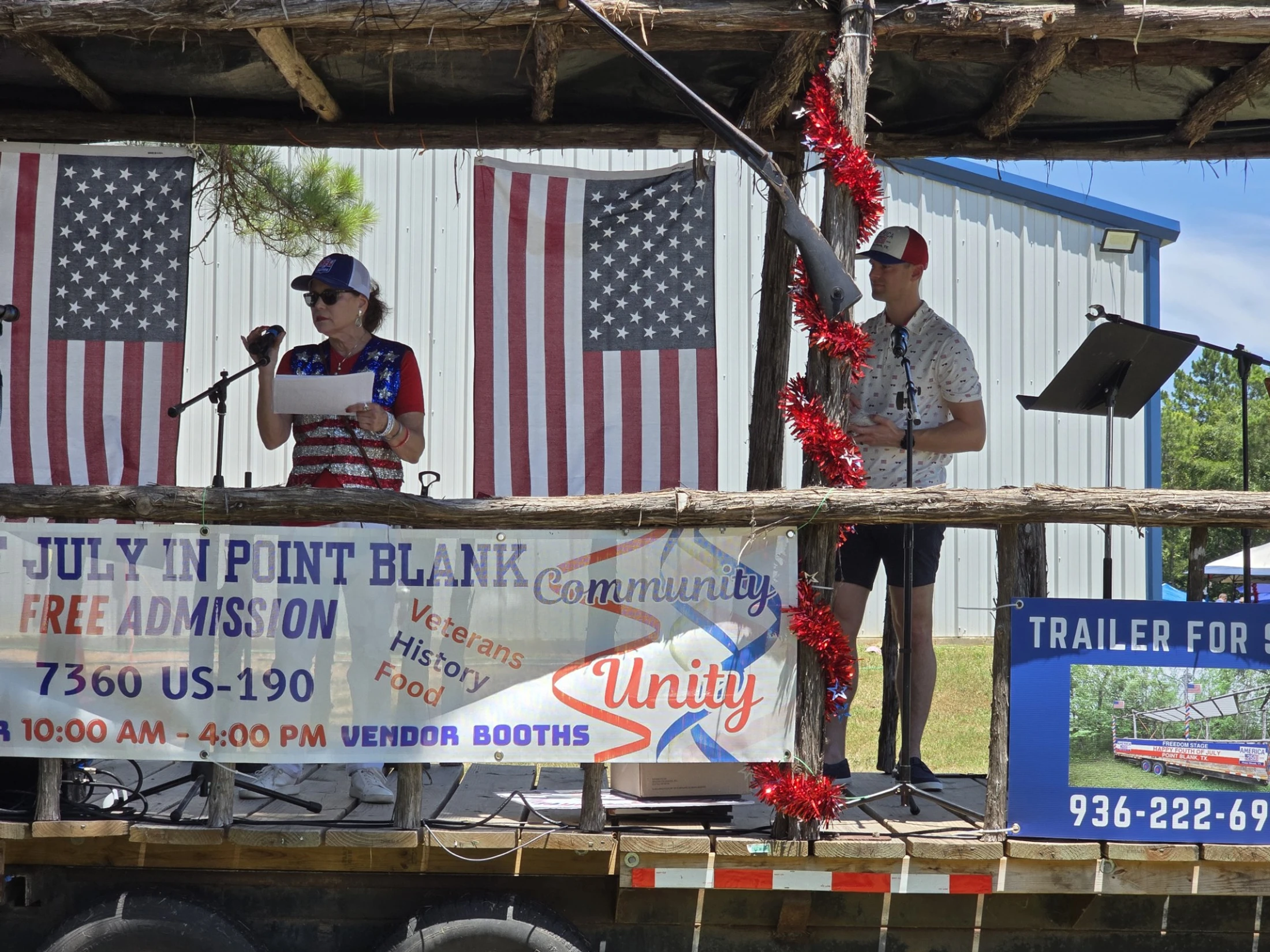 A woman and a man stand on a rustic wooden stage decorated with American flags and speak into microphones during an outdoor community festival in Point Blank.