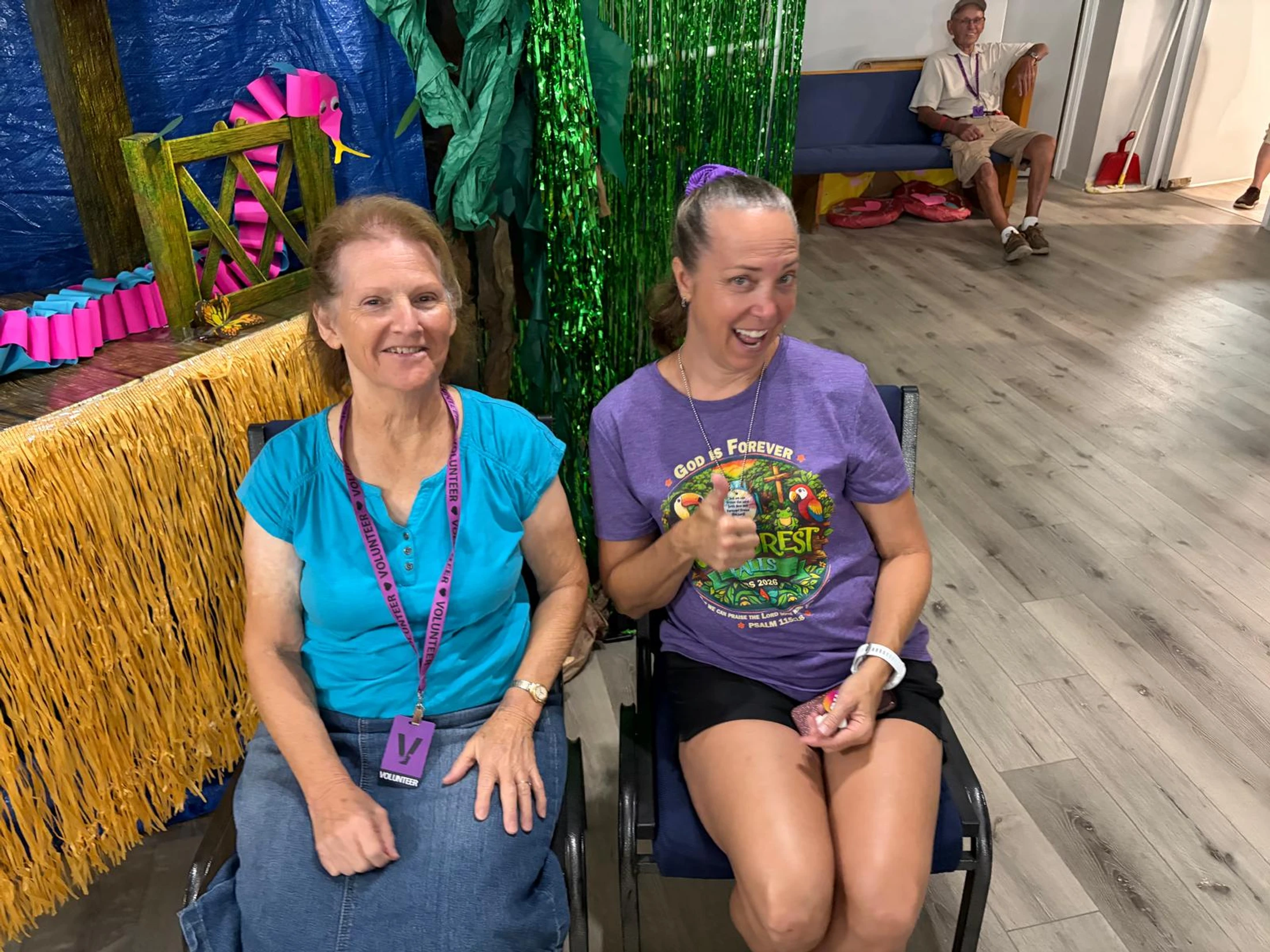 Two women wearing volunteer lanyards sit side-by-side and smile for the camera. The background features jungle-themed decorations.