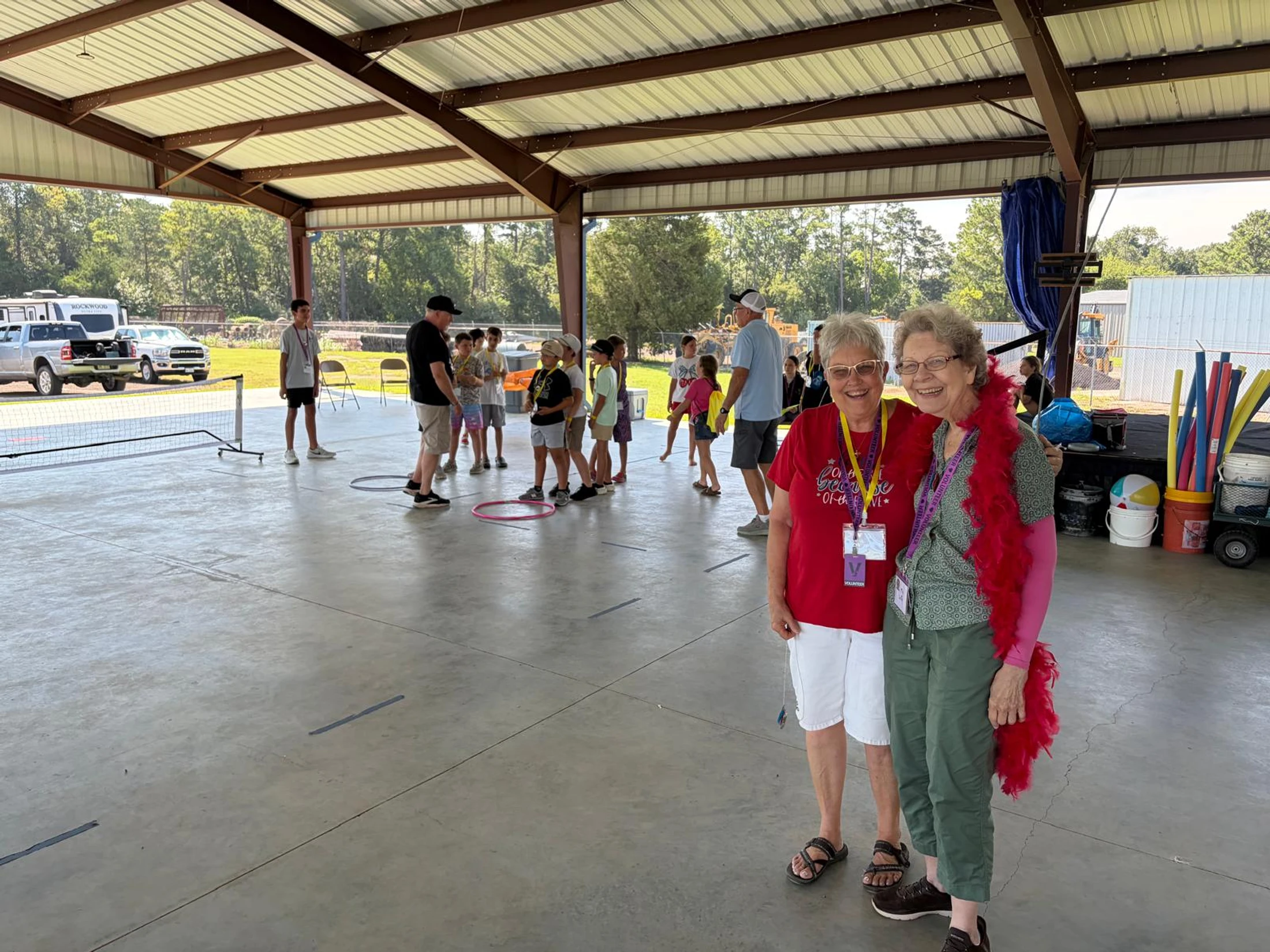 Two older women volunteers smile for the camera under a large pavilion, while children and other adults participate in activities in the background.