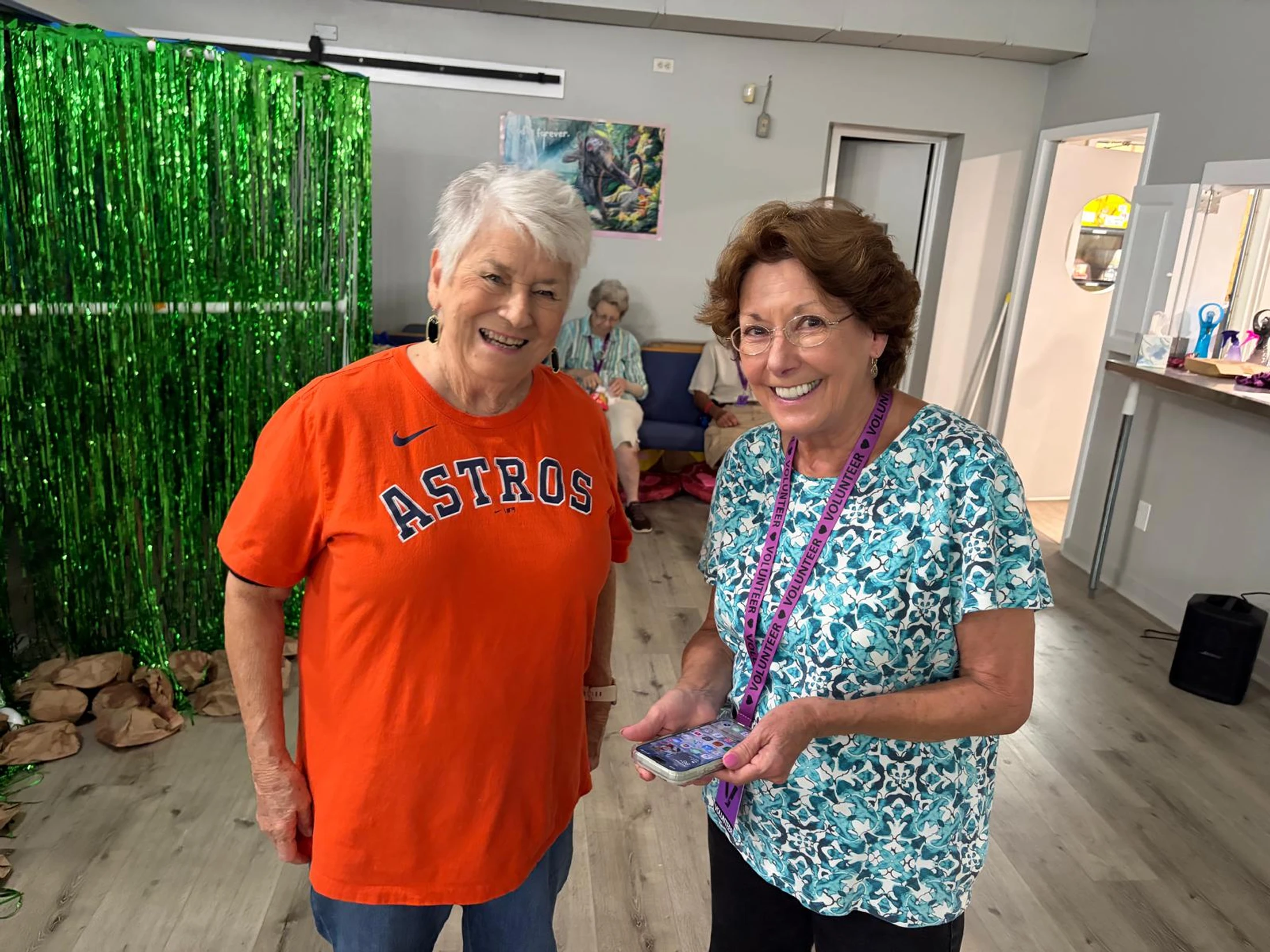 Two older women stand side-by-side smiling. The woman on the left wears an orange Astros shirt, and the woman on the right has on a volunteer lanyard.