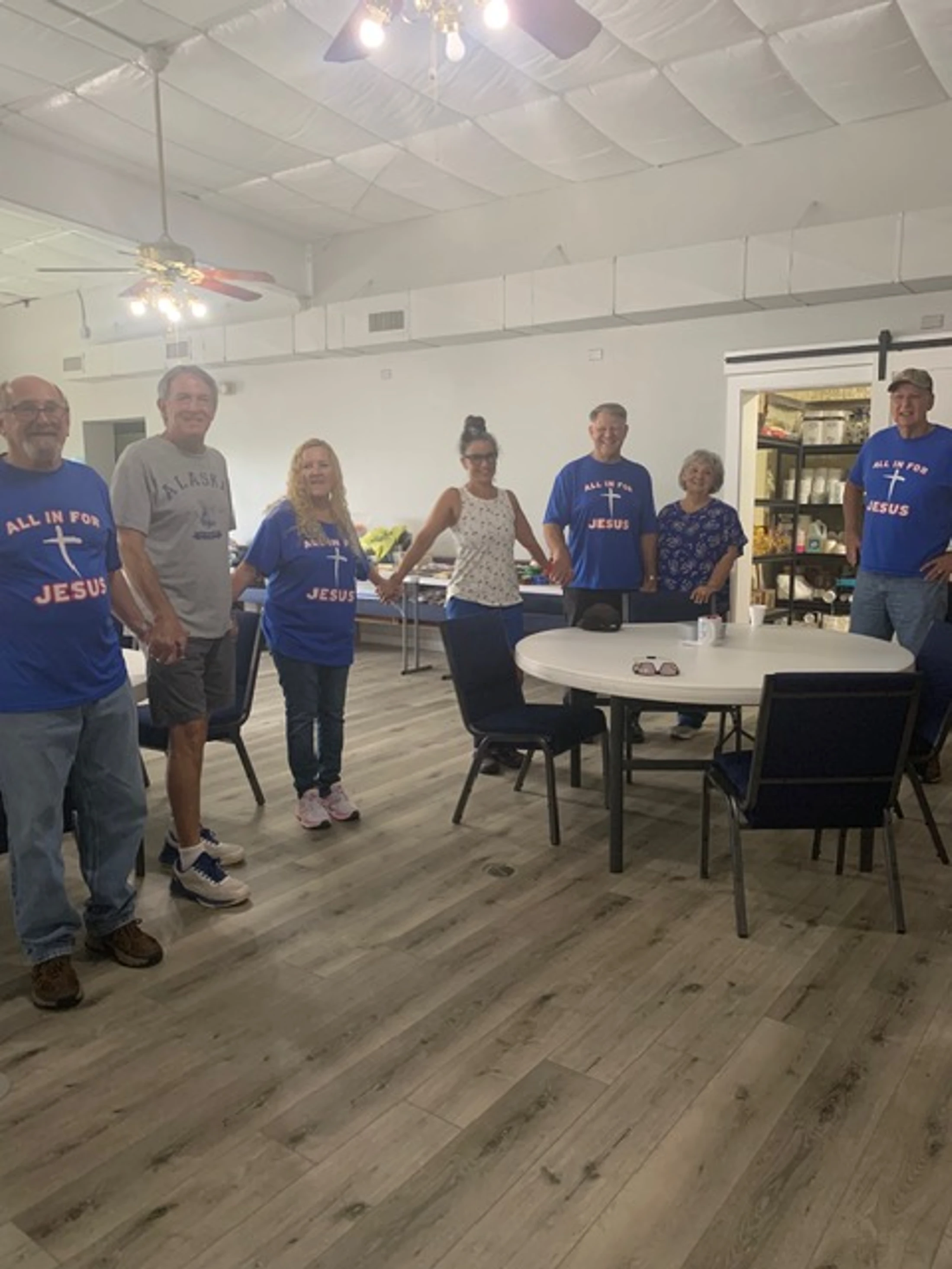 A group of seven adults stand in a room holding hands. Several wear matching blue t-shirts that say 'ALL IN FOR JESUS' with a white cross.