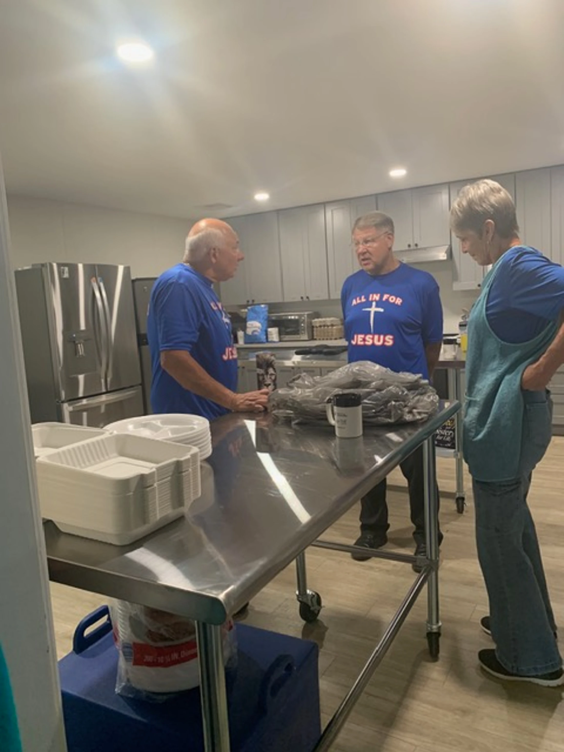 Three volunteers, two men and a woman, stand around a steel table in a kitchen. The men wear matching blue 'All In For Jesus' t-shirts.