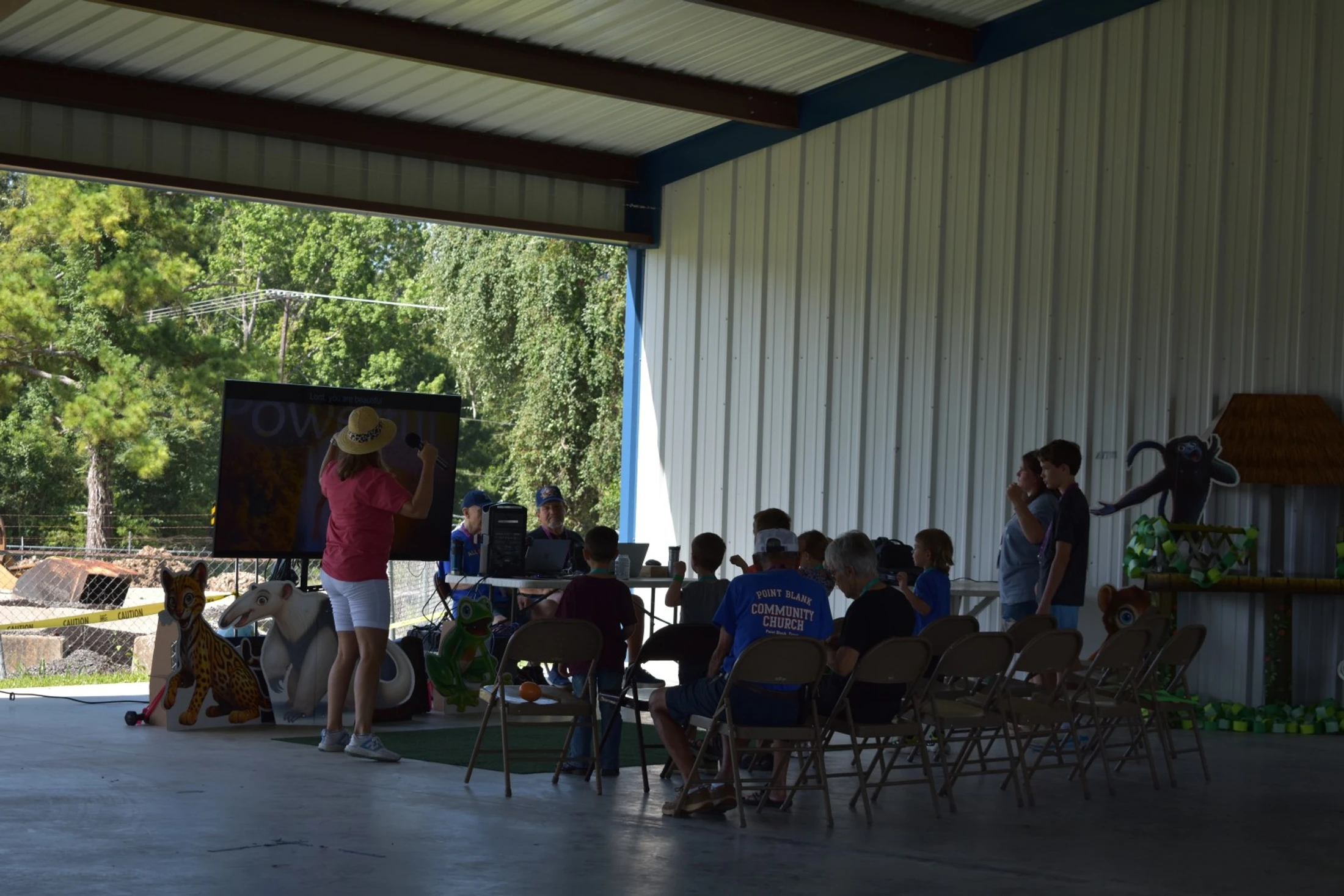 A woman in a pink shirt and straw hat speaks to an audience of children and adults seated in an open-air pavilion with animal decorations.