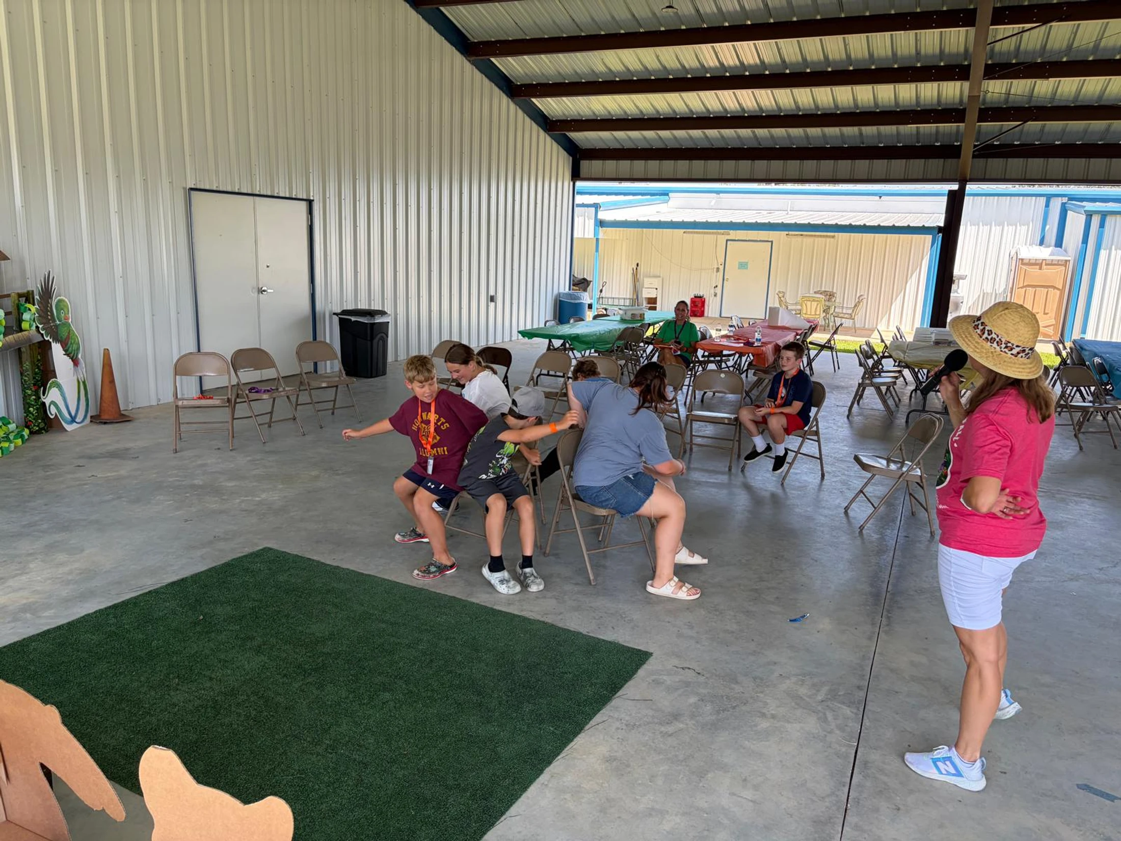 A group of children plays a game sitting on each other's laps in a line of chairs, while an adult supervisor watches in a large, open-air pavilion.