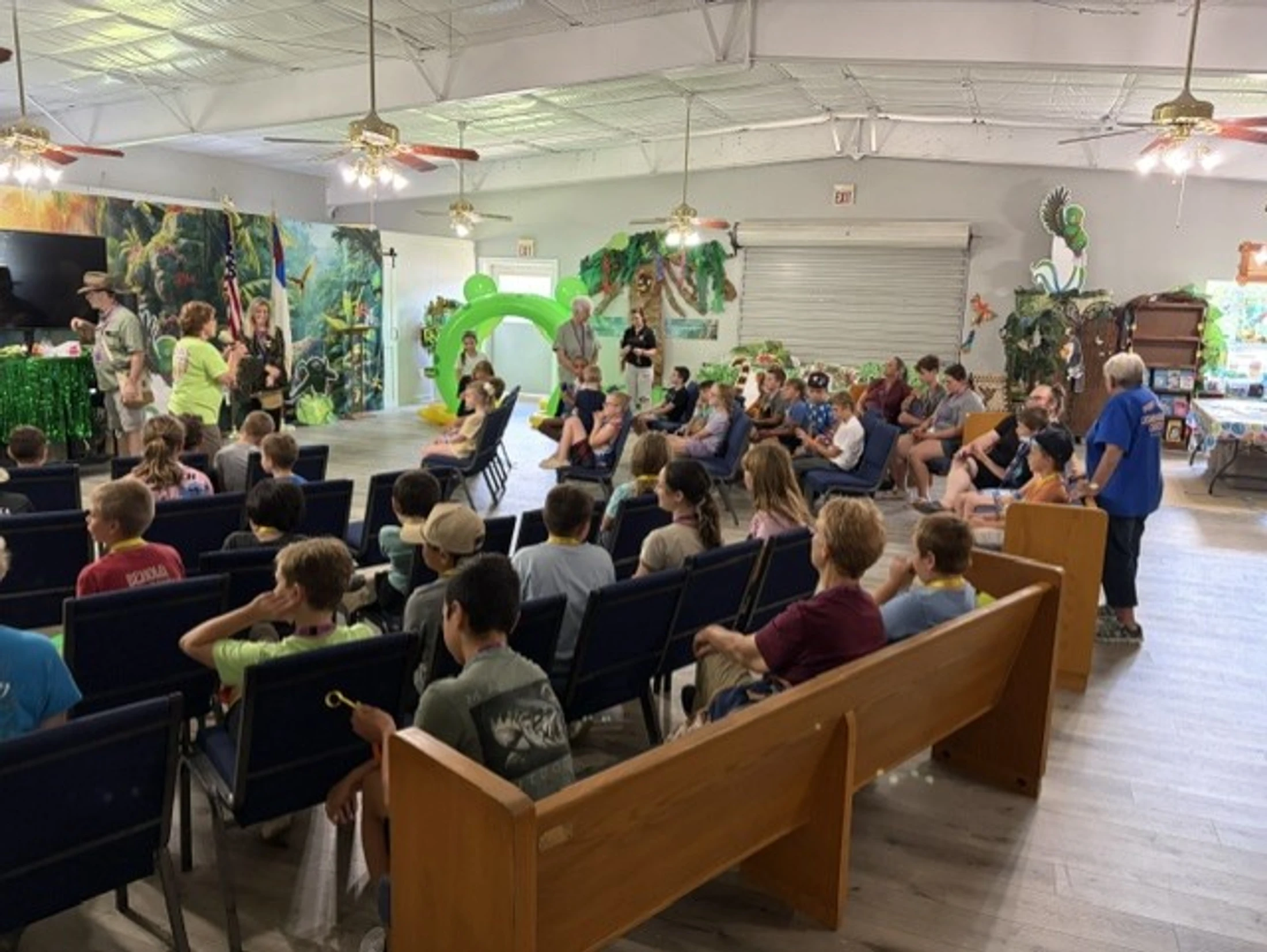 A large group of children sit in chairs and pews, attentively watching a presentation in a community hall decorated with a jungle and safari theme.