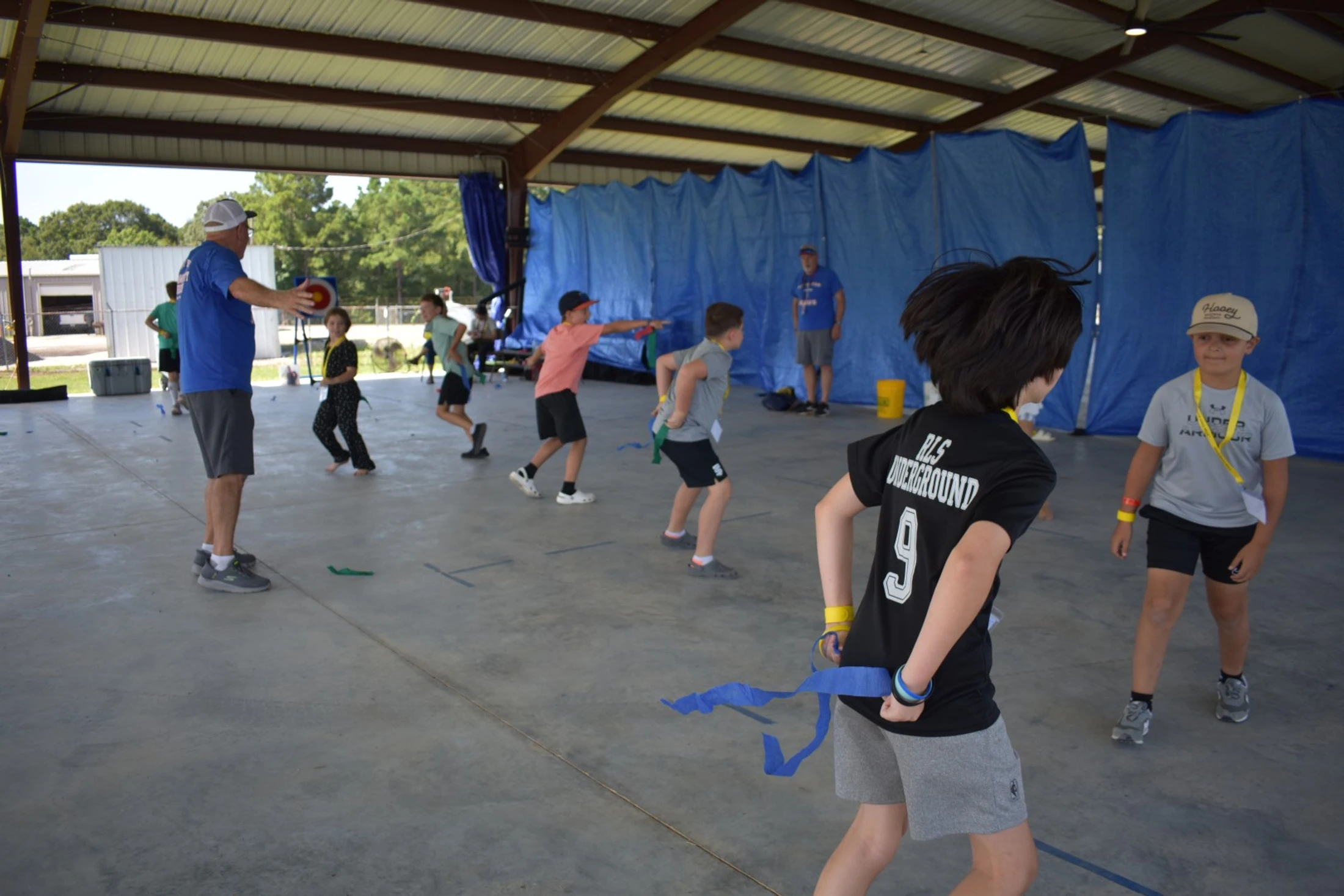 A group of children and an adult leader play a game, possibly flag football, under an open-air pavilion with a concrete floor.