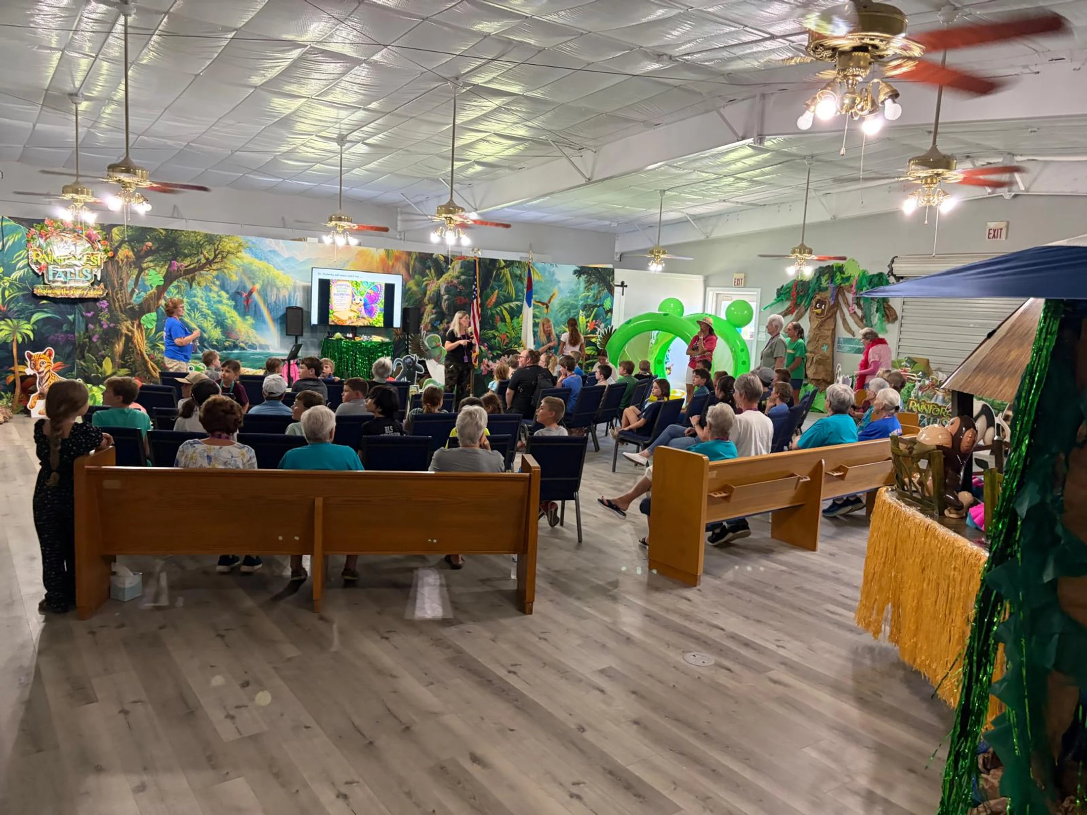 A large group of children and adults seated in a hall decorated with a vibrant rainforest theme. Two women lead the assembly from a stage at the front.