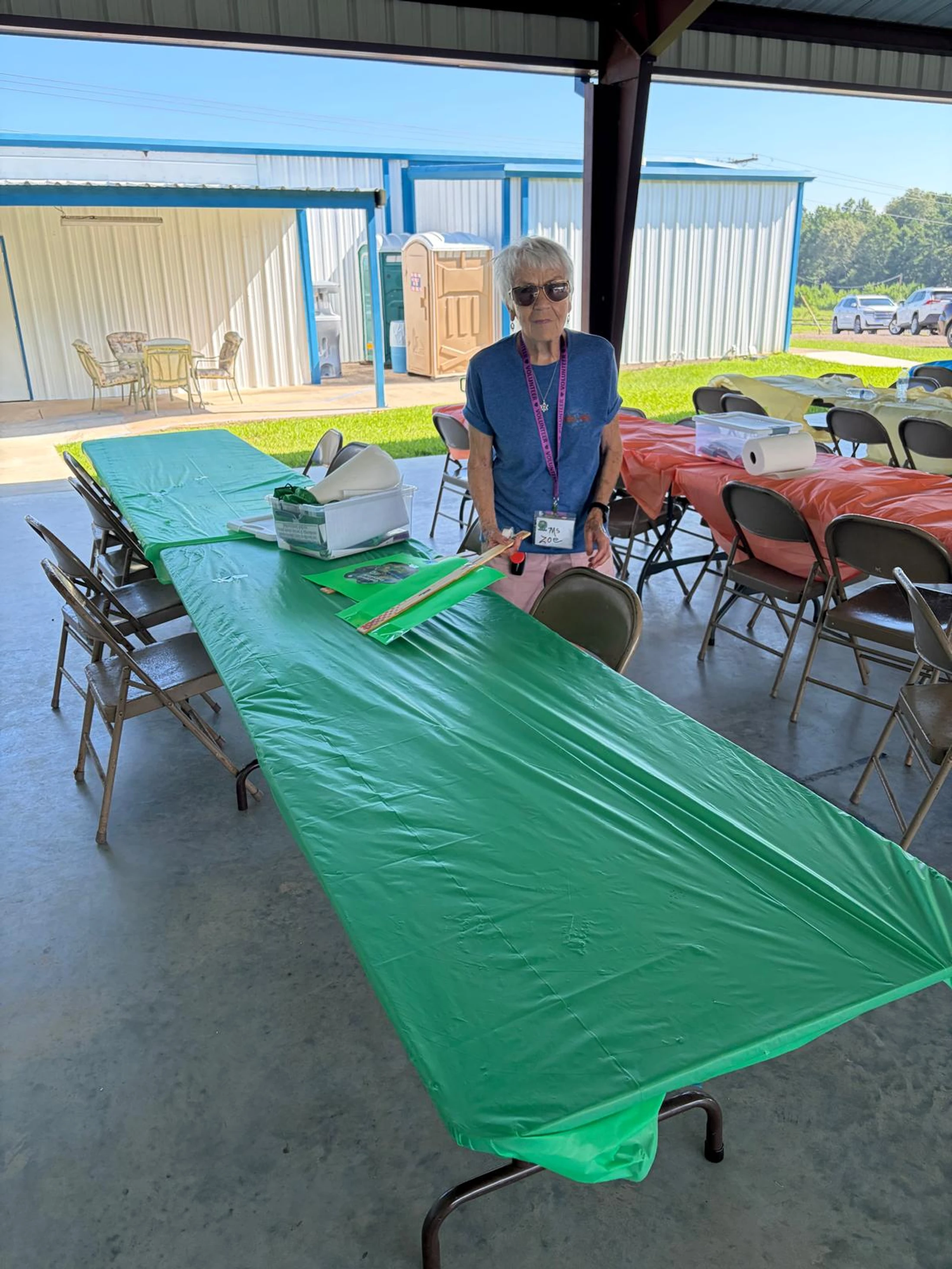 A woman with short gray hair and sunglasses stands behind a long table with a green tablecloth at an outdoor event. She wears a volunteer lanyard.