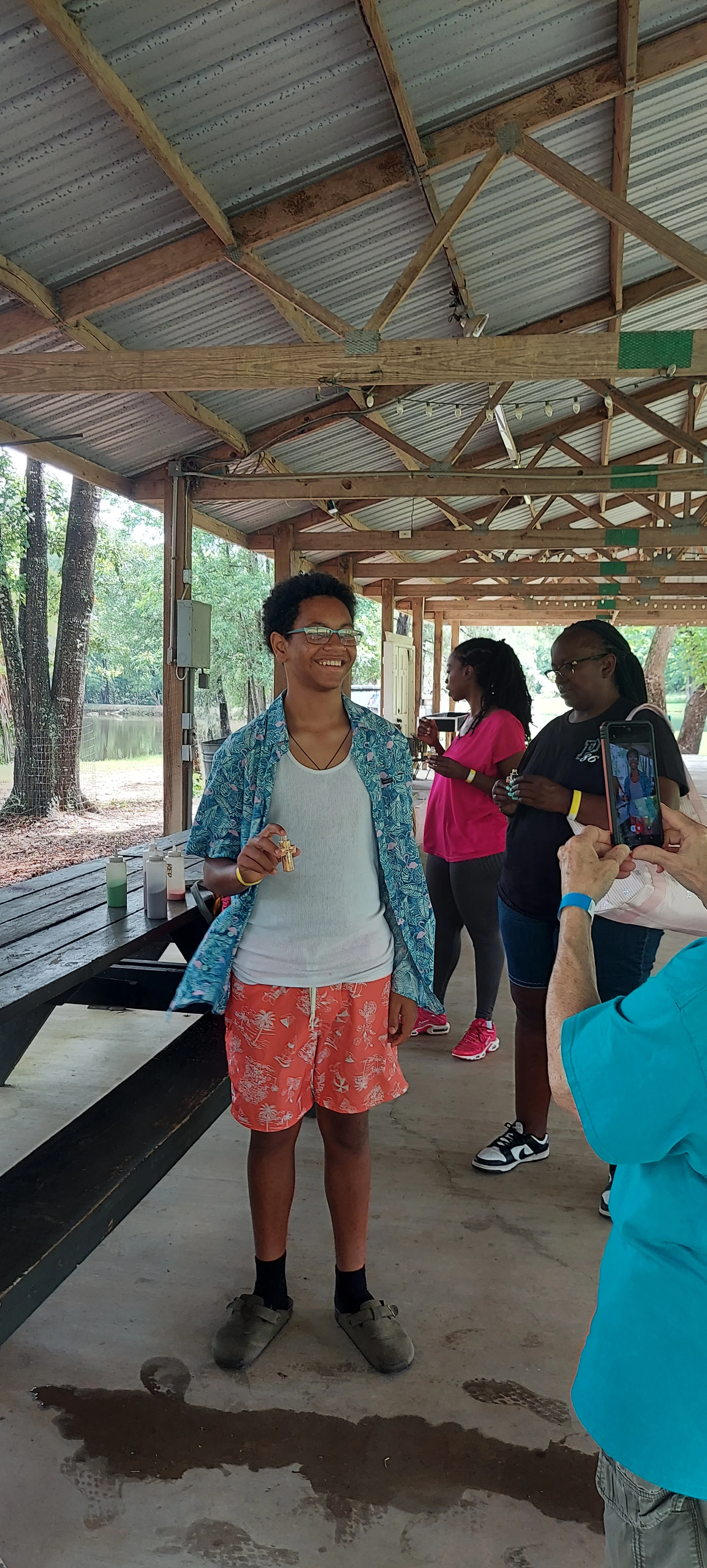 A young person with glasses smiles while posing for a photo under a park pavilion. They wear a patterned shirt and orange shorts and hold a small golden cross.