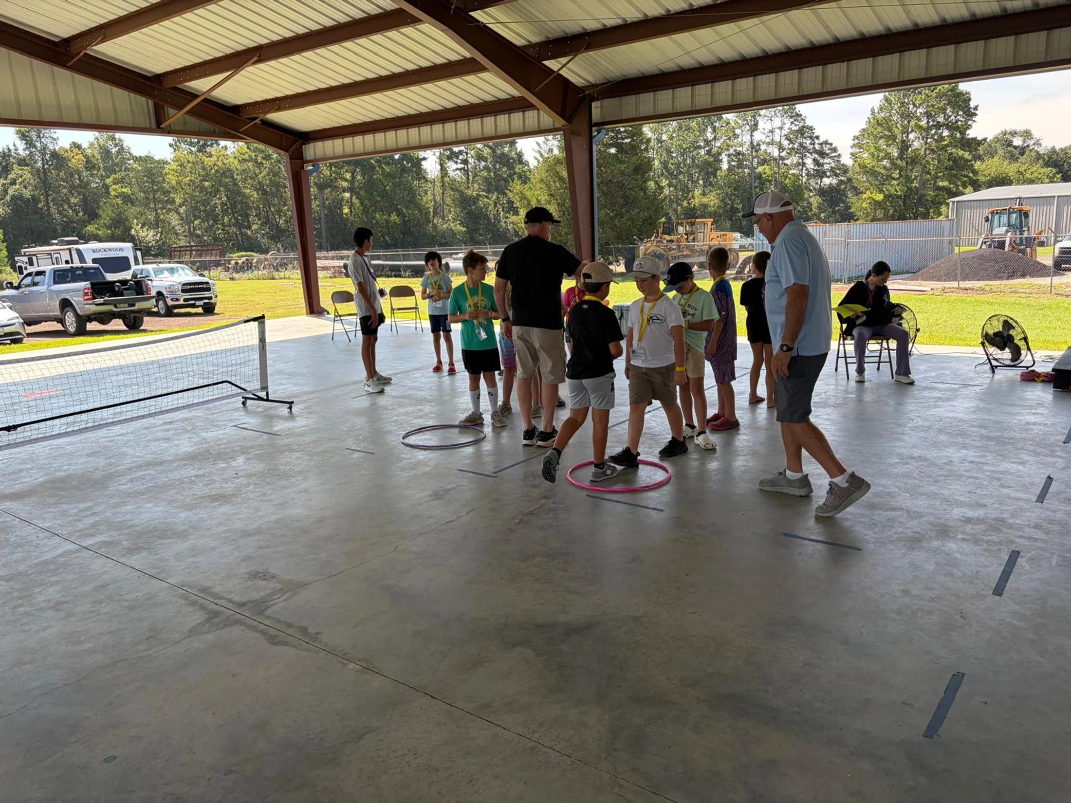 A group of young boys and adult leaders stand in a circle under a large metal pavilion, participating in a group activity with hula hoops on the concrete floor.