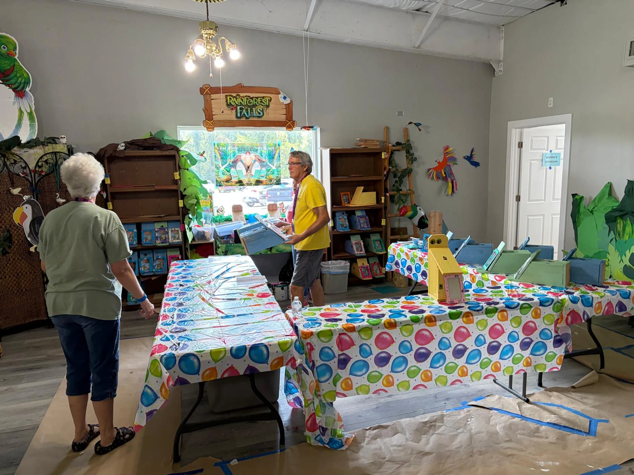 A man and a woman set up a room with a "Rainforest Falls" theme, preparing crafts on tables covered with colorful balloon-patterned tablecloths.