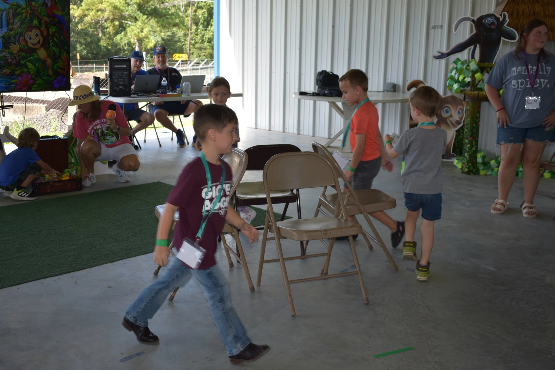 A group of young boys walks around metal folding chairs during a game at a jungle-themed event, with adults supervising in the background.