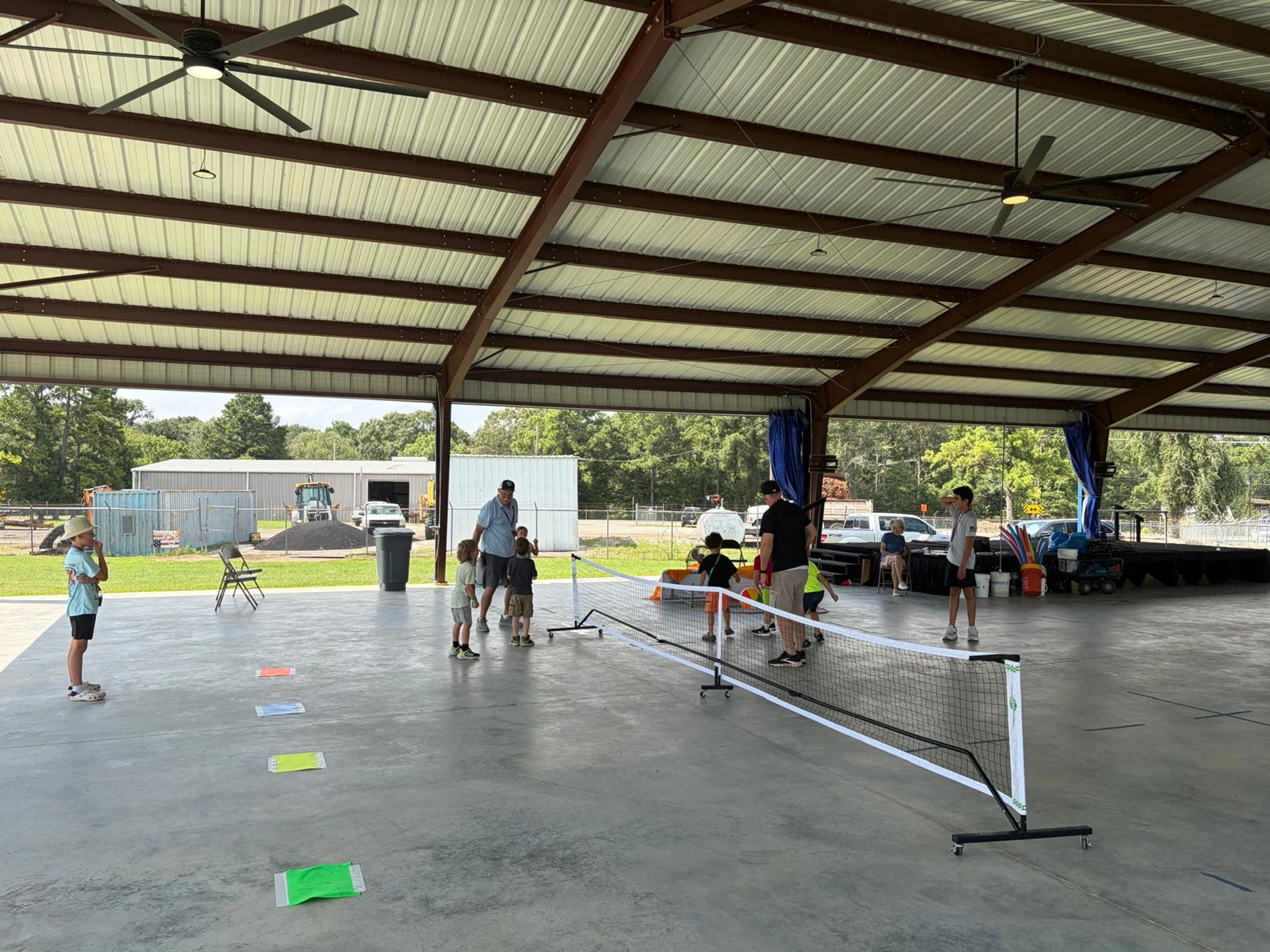 A group of children and adults are gathered around a small portable net on a concrete court under a large metal pavilion for a sports lesson.