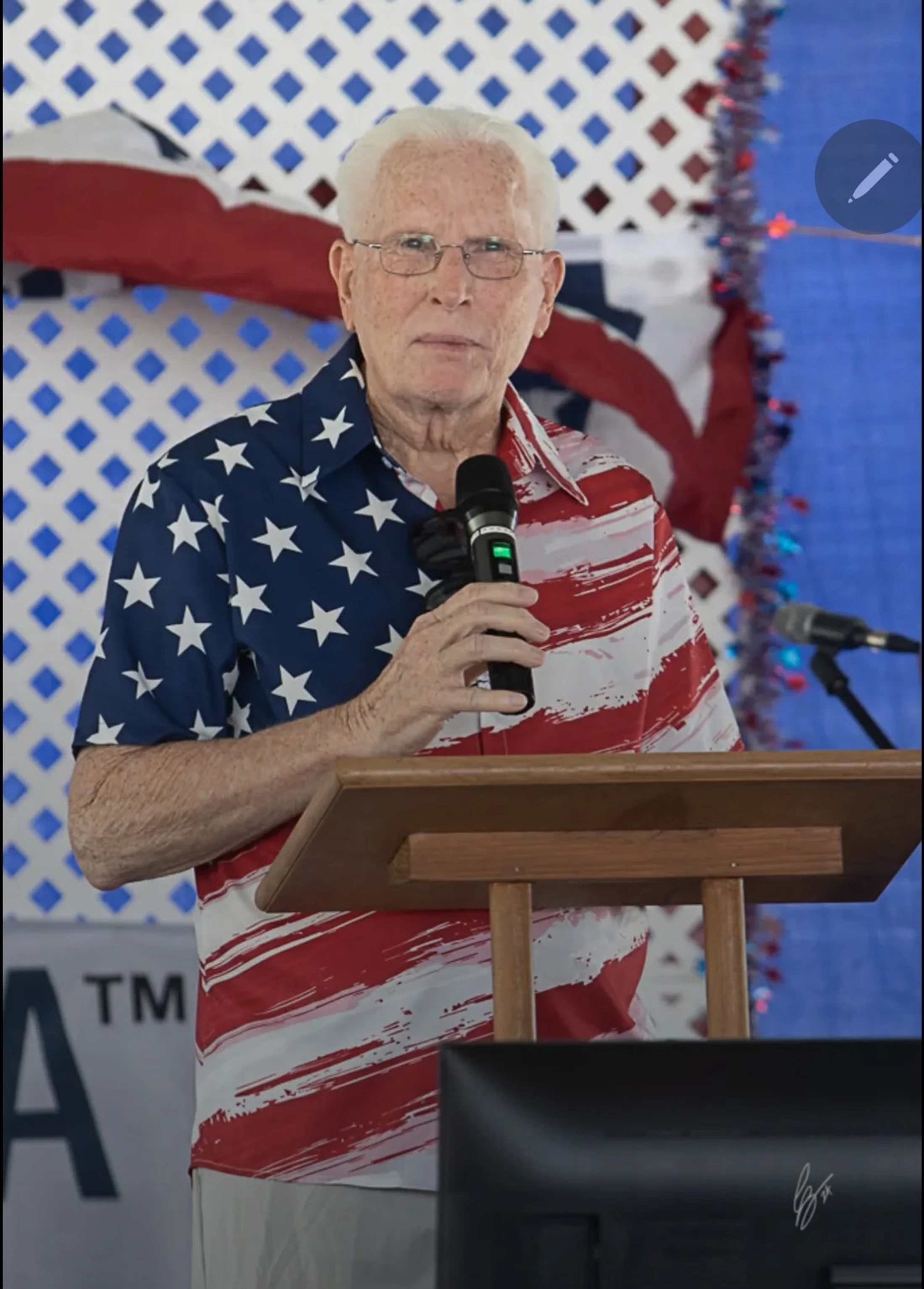 An older man with white hair and glasses, wearing a patriotic US flag shirt, stands at a lectern holding a microphone and addressing an audience.