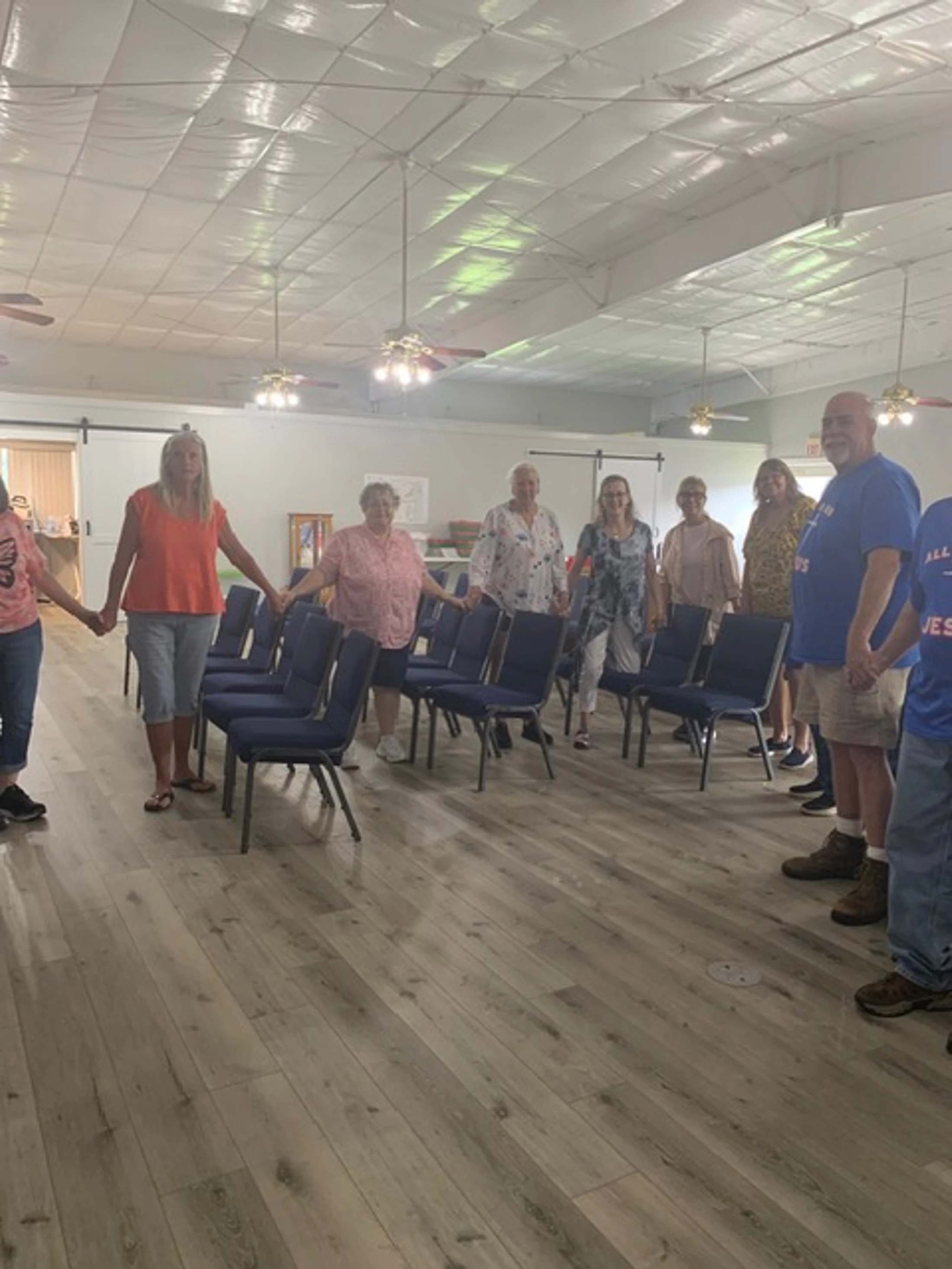 A group of adults stand in a large circle holding hands in a community hall. In the middle of the circle are rows of empty blue chairs.