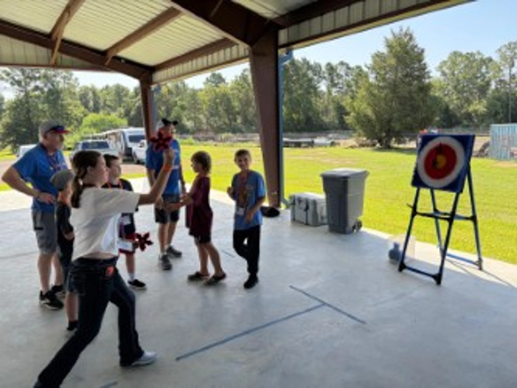 A group of children and adults under a pavilion play a target game. A young girl in a white shirt is throwing an object at a colorful target on a stand.