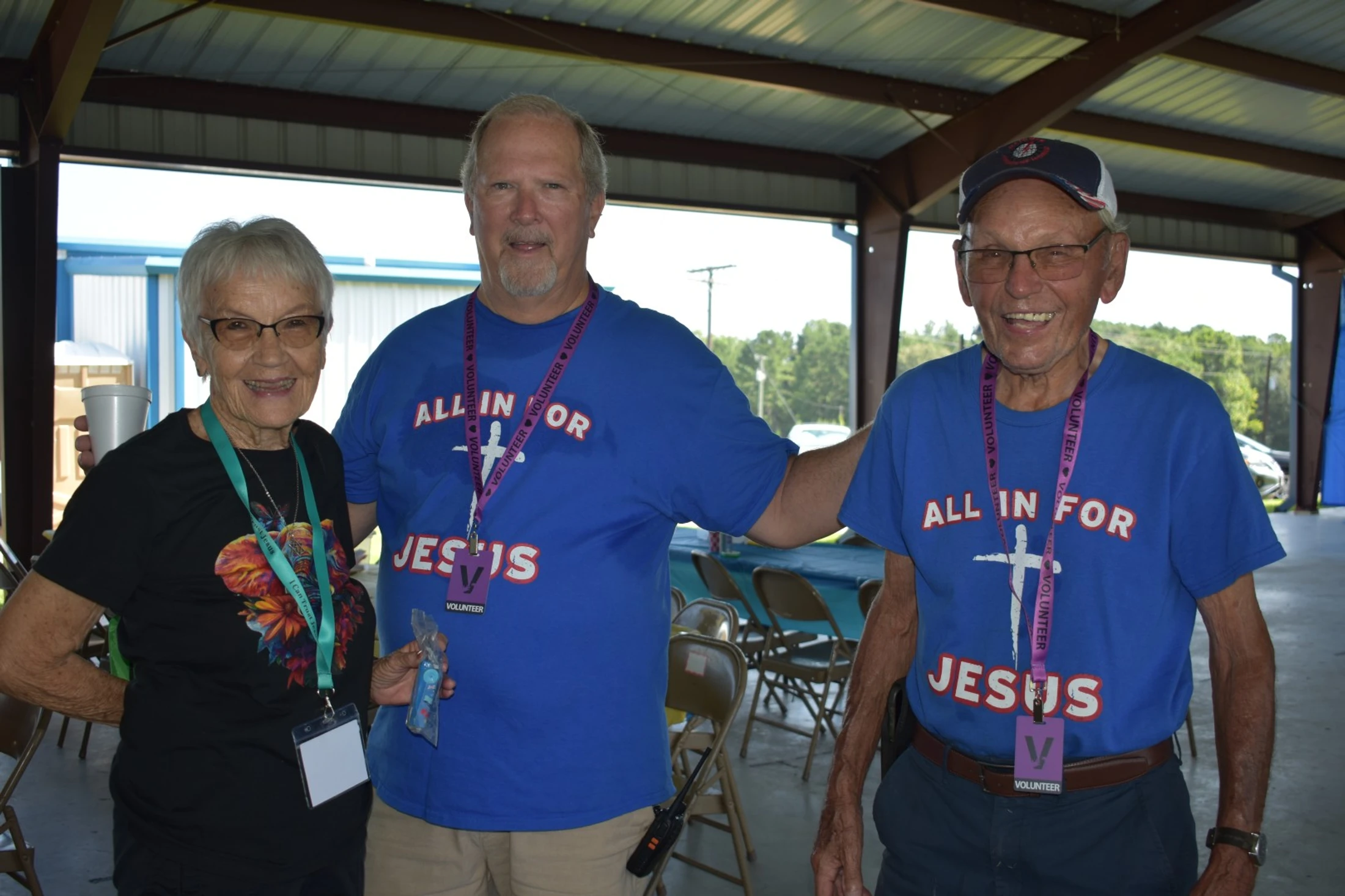 Three volunteers, an older woman and two men, smile for the camera at an event. The men wear matching blue t-shirts that say 'All in for Jesus'.