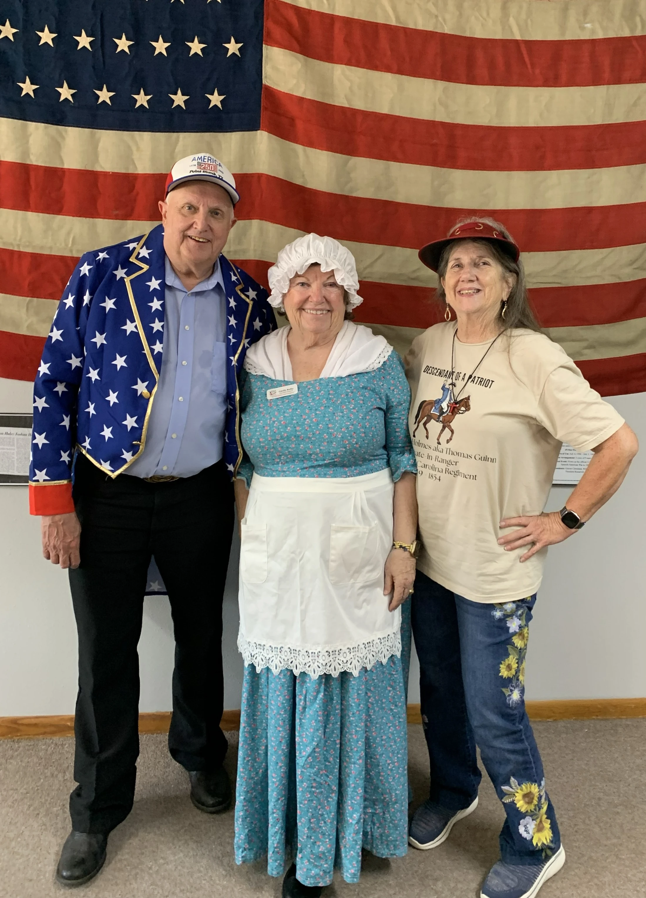 A man and two women in patriotic and colonial-style costumes smile while standing in front of a large American flag.