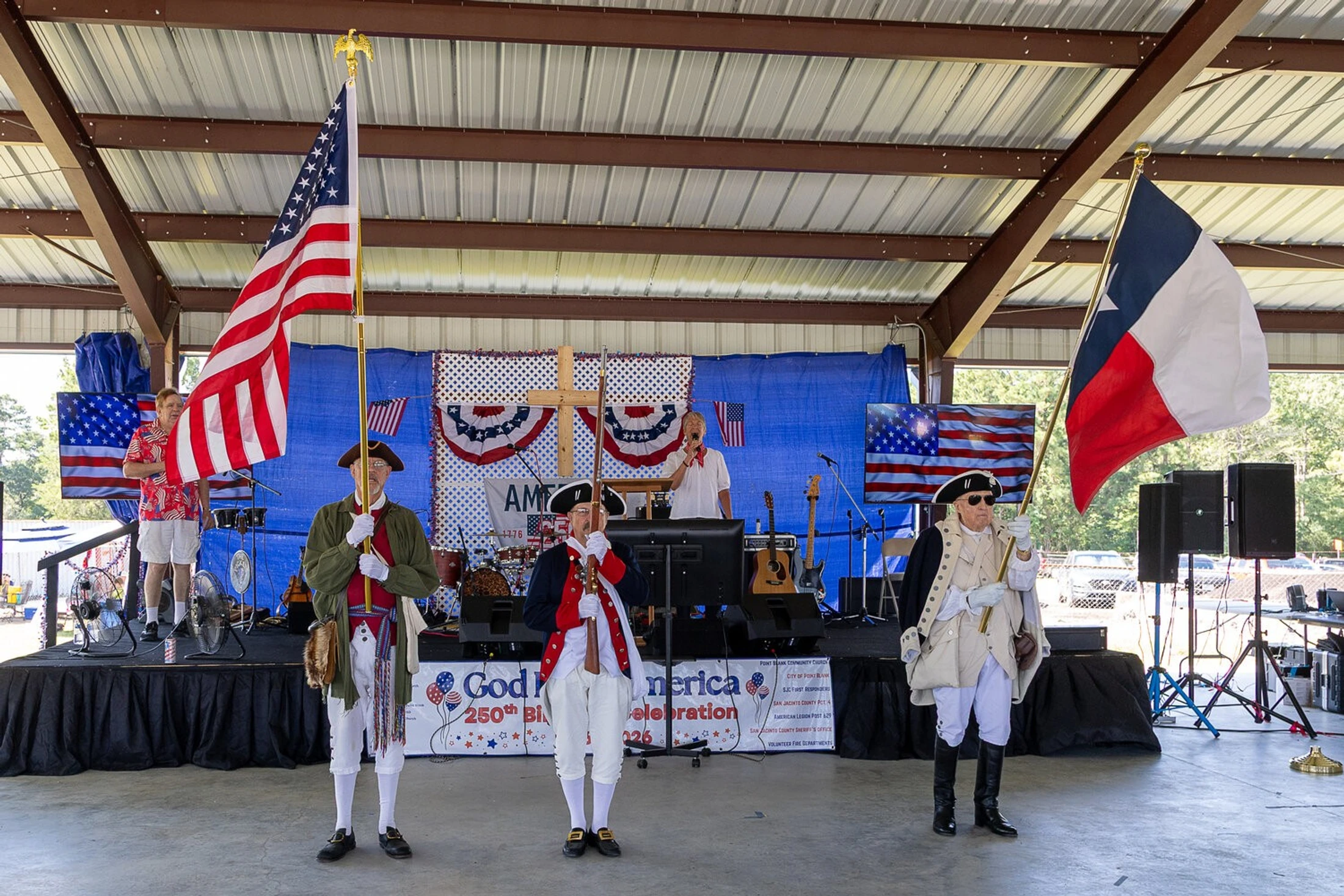 Three men in colonial-era costumes present the American and Texas flags on a stage decorated with patriotic bunting during an outdoor event.