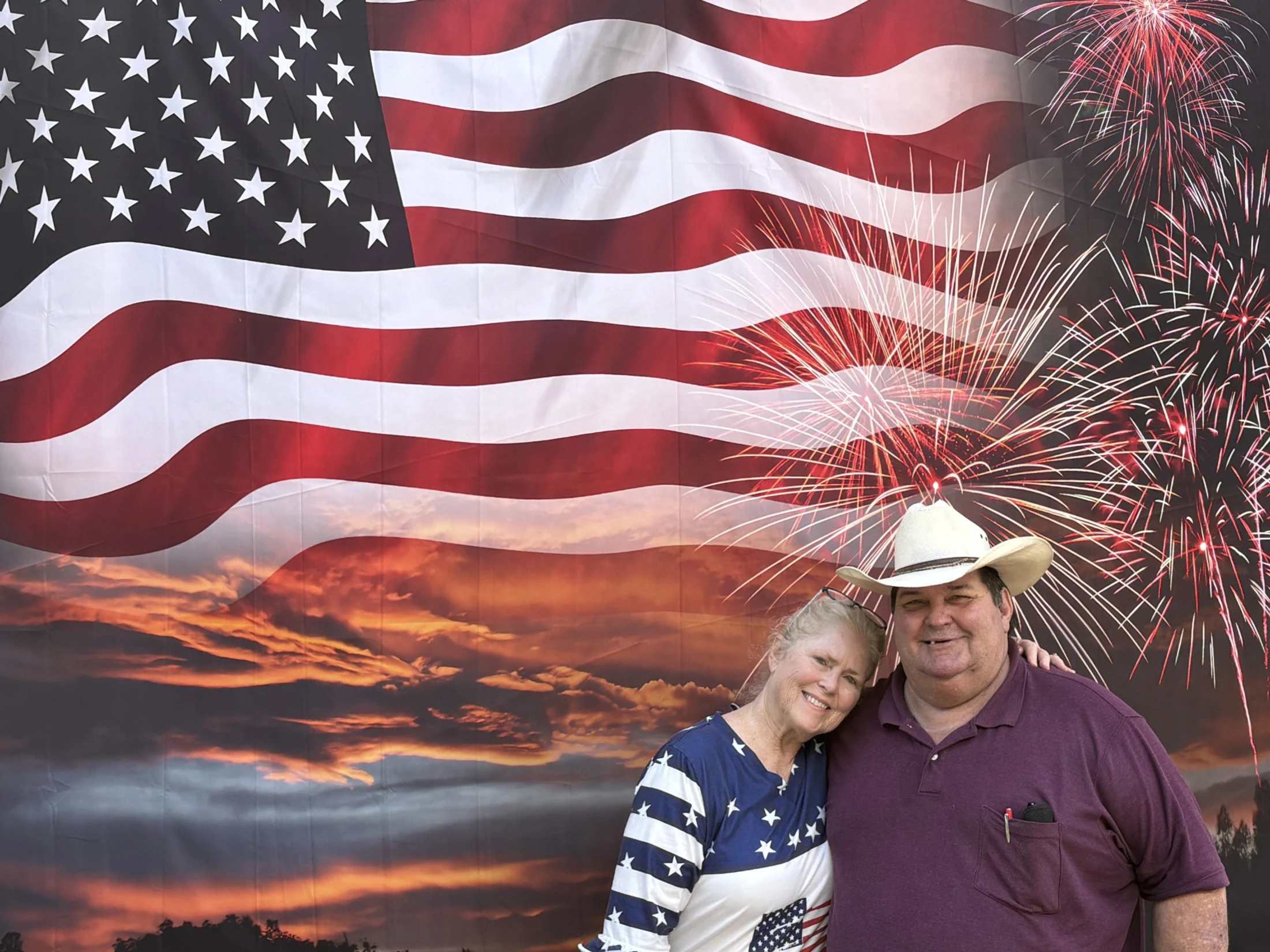 A smiling couple poses in front of a patriotic backdrop with an American flag, a sunset, and fireworks. The man wears a cowboy hat.