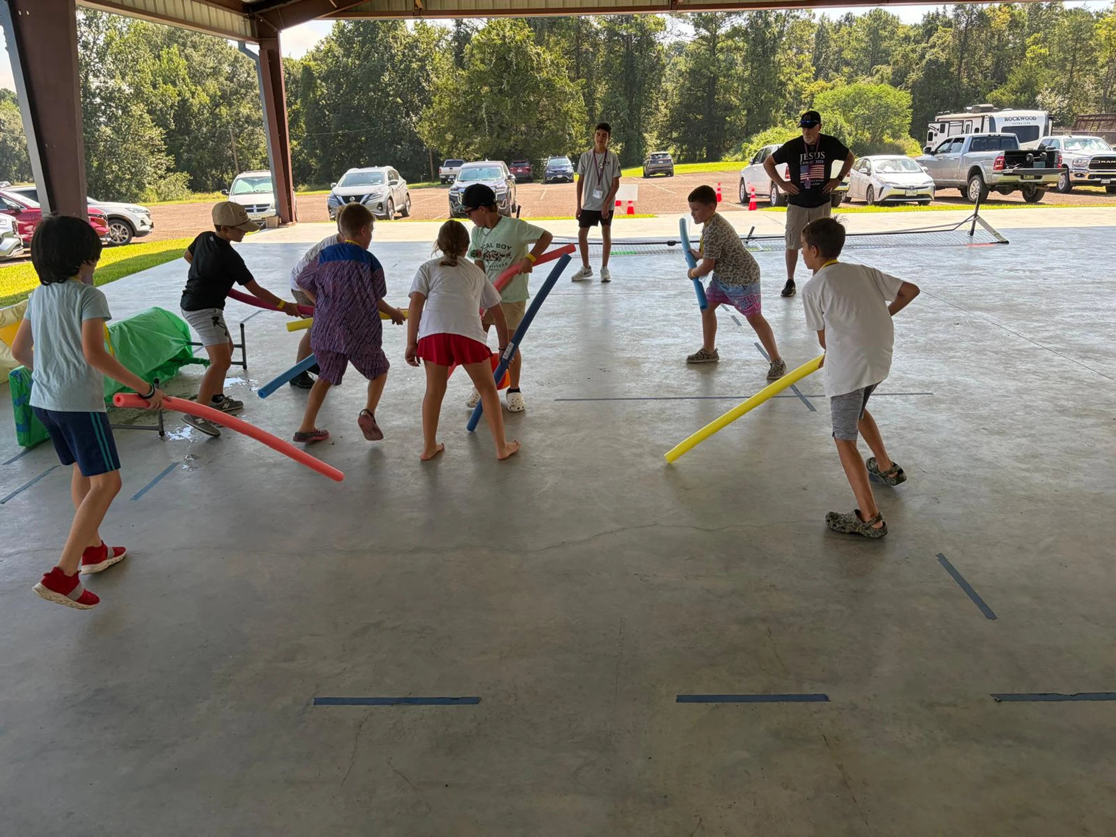 A group of children play fighting with colorful pool noodles under a large outdoor pavilion while two adults supervise in the background.