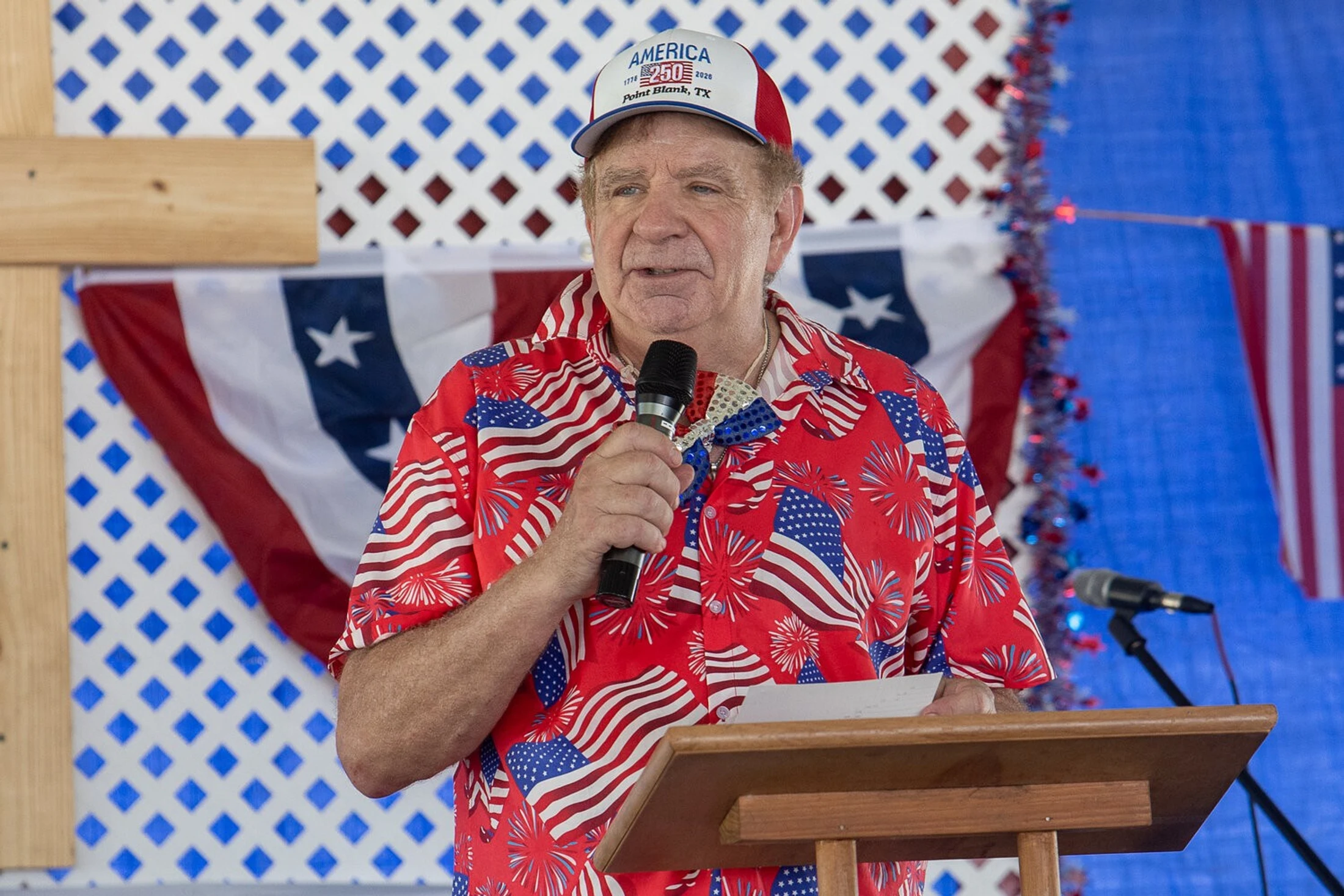An older man in a red, white, and blue shirt and an "America 250" hat speaks into a microphone while standing at a wooden podium.