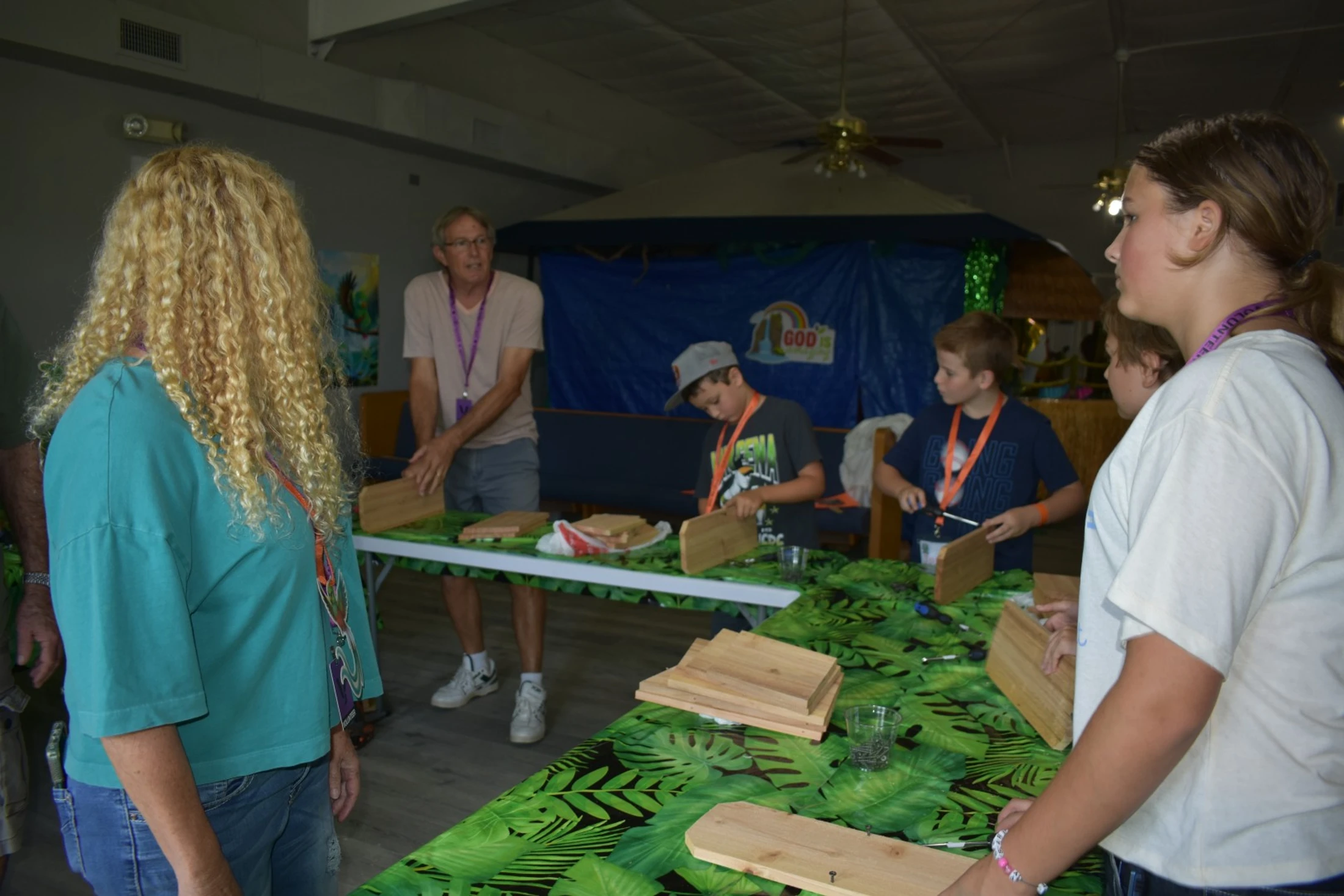 Children and adults stand around a leaf-patterned table, assembling small wooden projects with screws and tools while an instructor gives guidance.