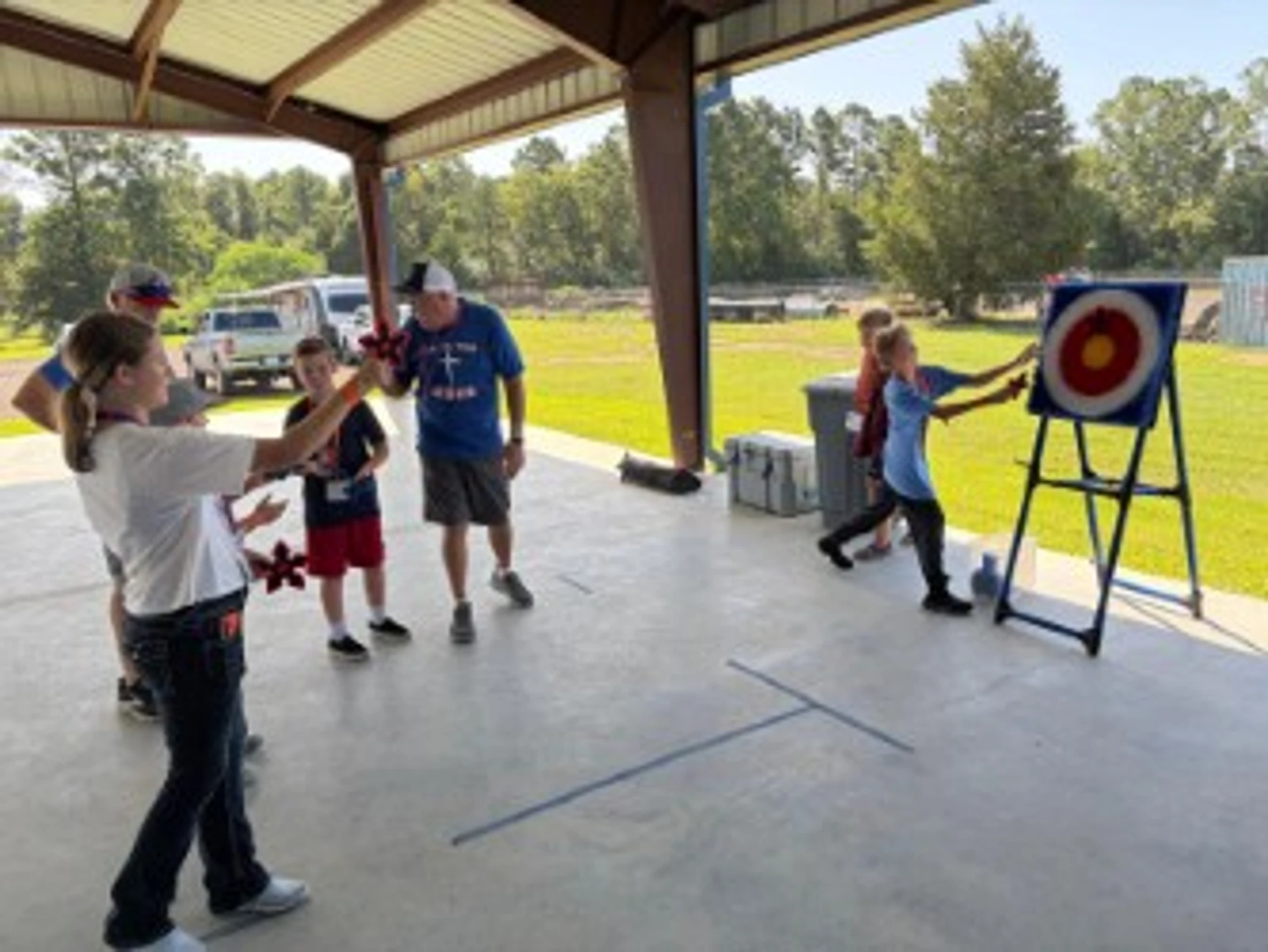 A group of children and adults under a covered pavilion play a game. A young girl prepares to throw a red star at a circular target held up by a stand.