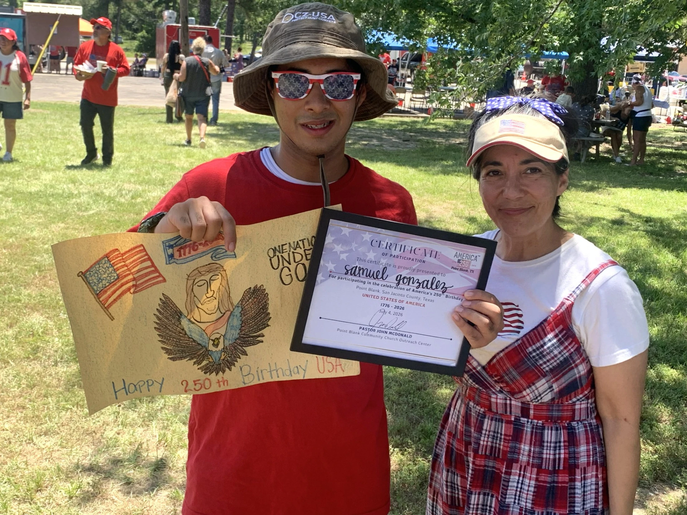 A young man and a woman proudly display a patriotic poster and a framed certificate at an outdoor community celebration for America's 250th birthday.