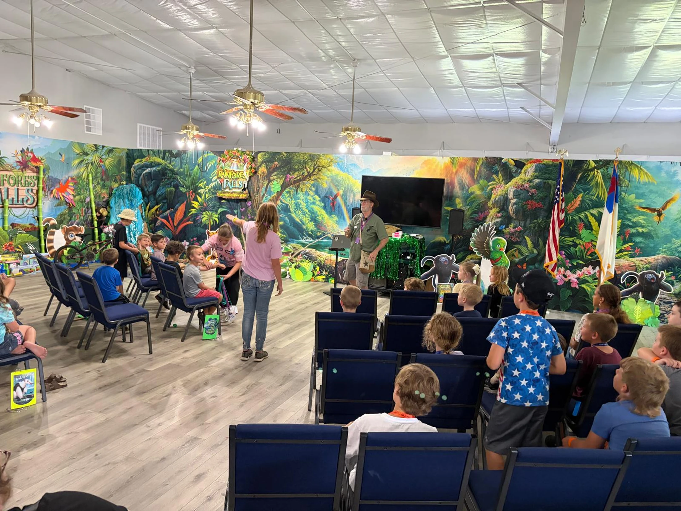 A group of children sit in chairs watching a presentation in a room decorated with a colorful, jungle-themed mural that says 'Rainforest Falls'.