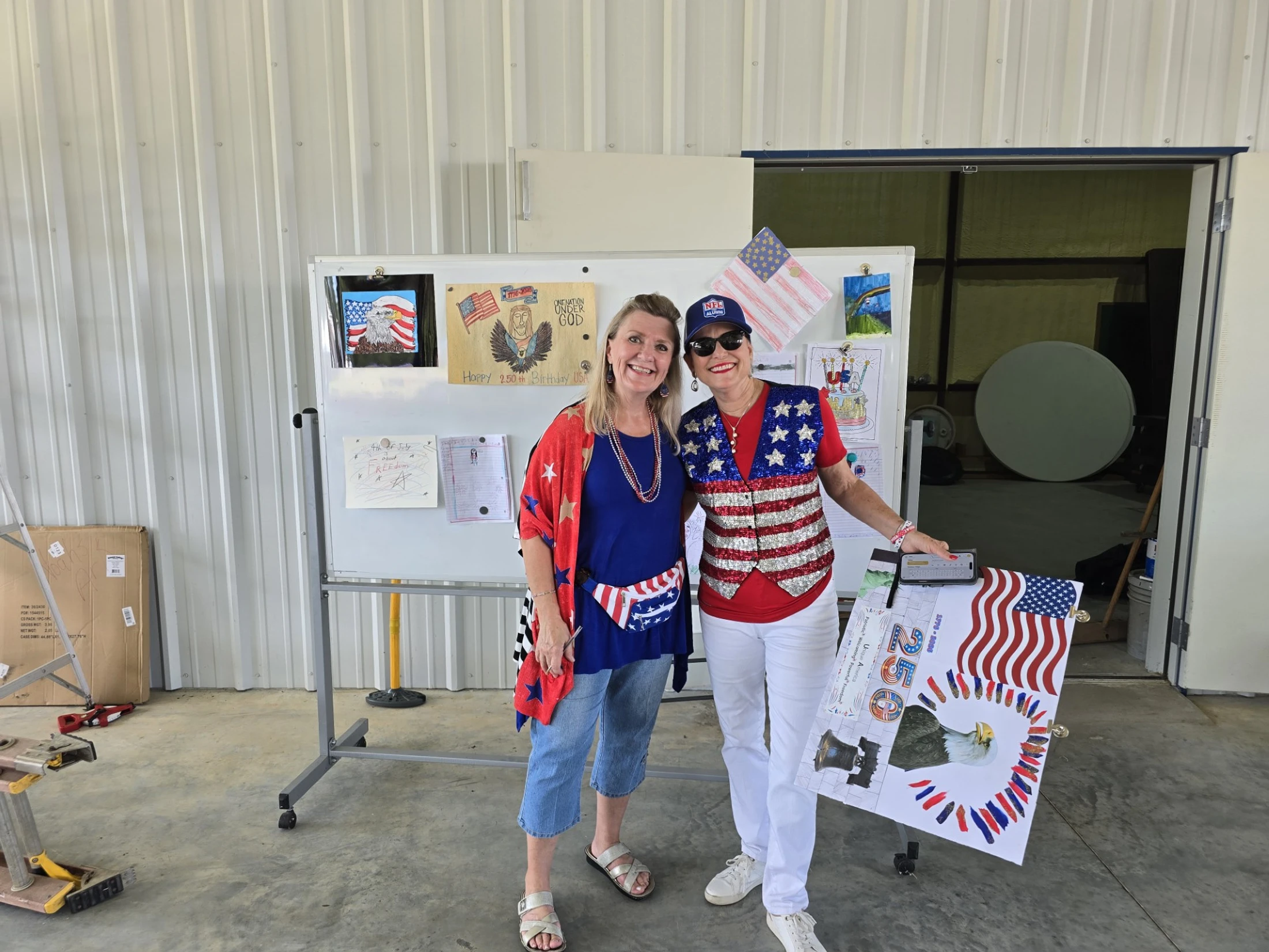 Two smiling women in red, white, and blue outfits stand in front of a whiteboard displaying patriotic children's art in a garage setting.