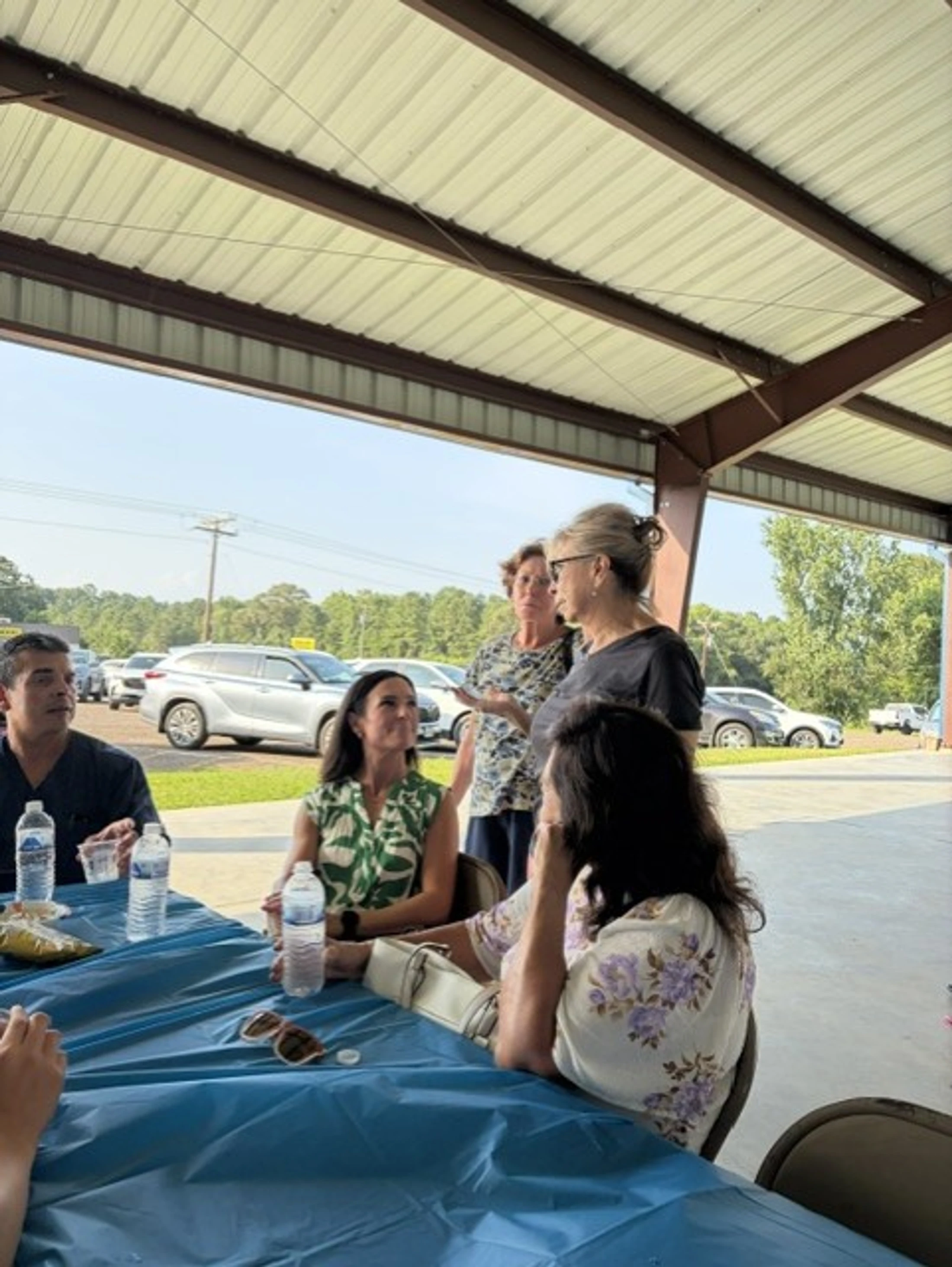 A group of people gather around a table with a blue tablecloth under a pavilion, engaged in conversation on a sunny day with cars parked in the background.