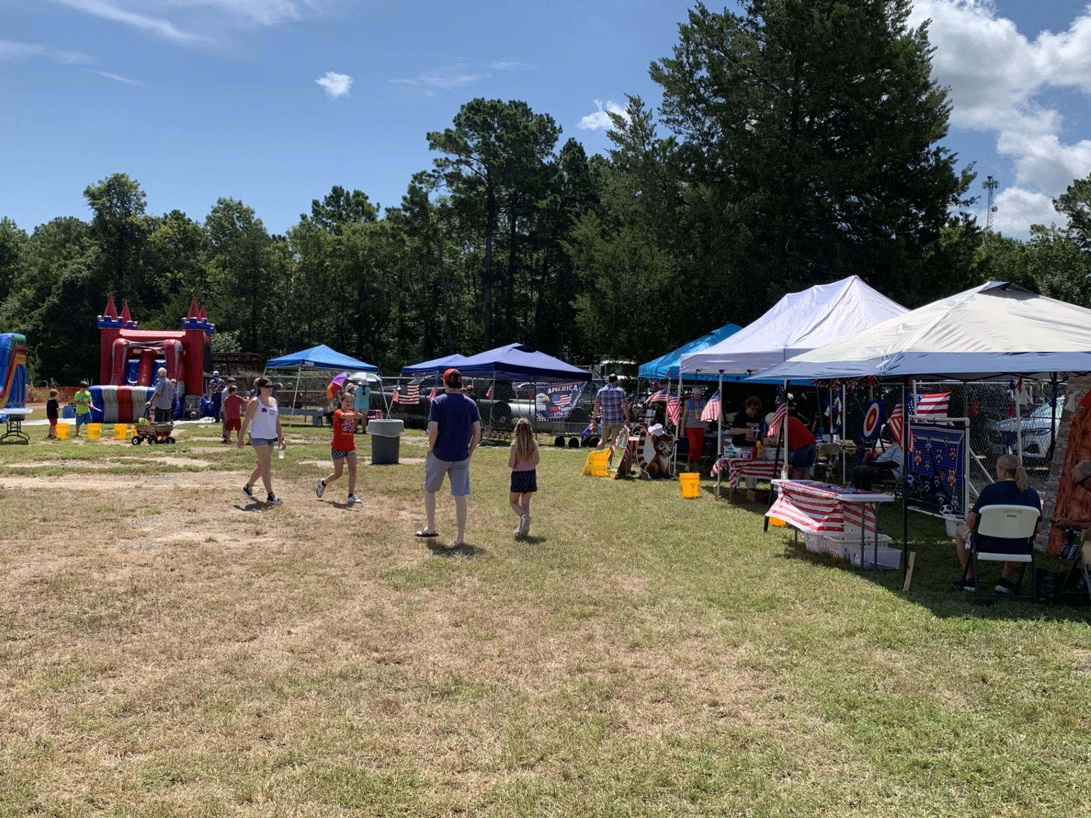 People walk through a grassy field at an outdoor festival with vendor tents decorated with American flags and an inflatable bounce house in the background.