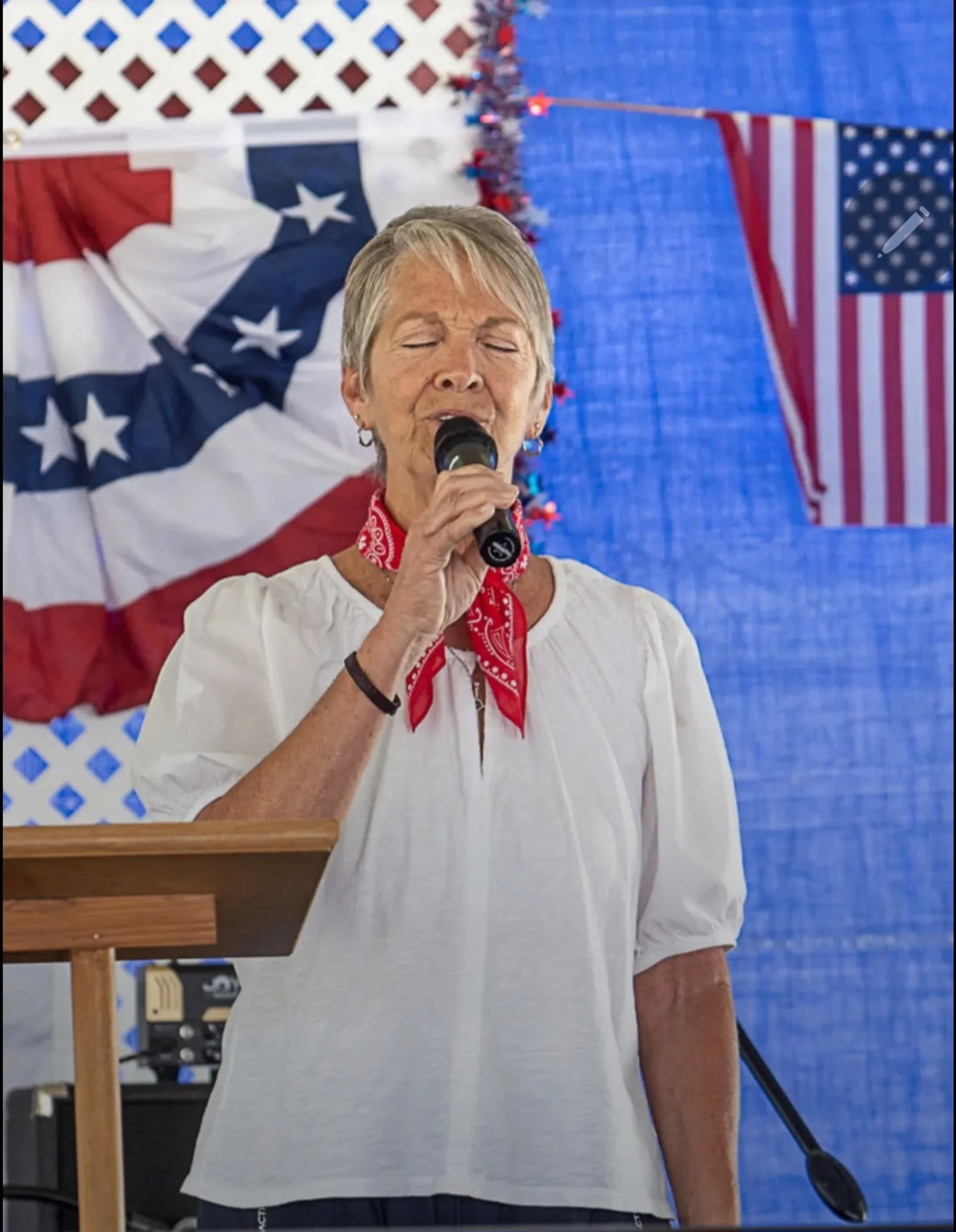 A woman with short gray hair and a red bandana sings into a microphone with her eyes closed. The background is decorated with American flags.
