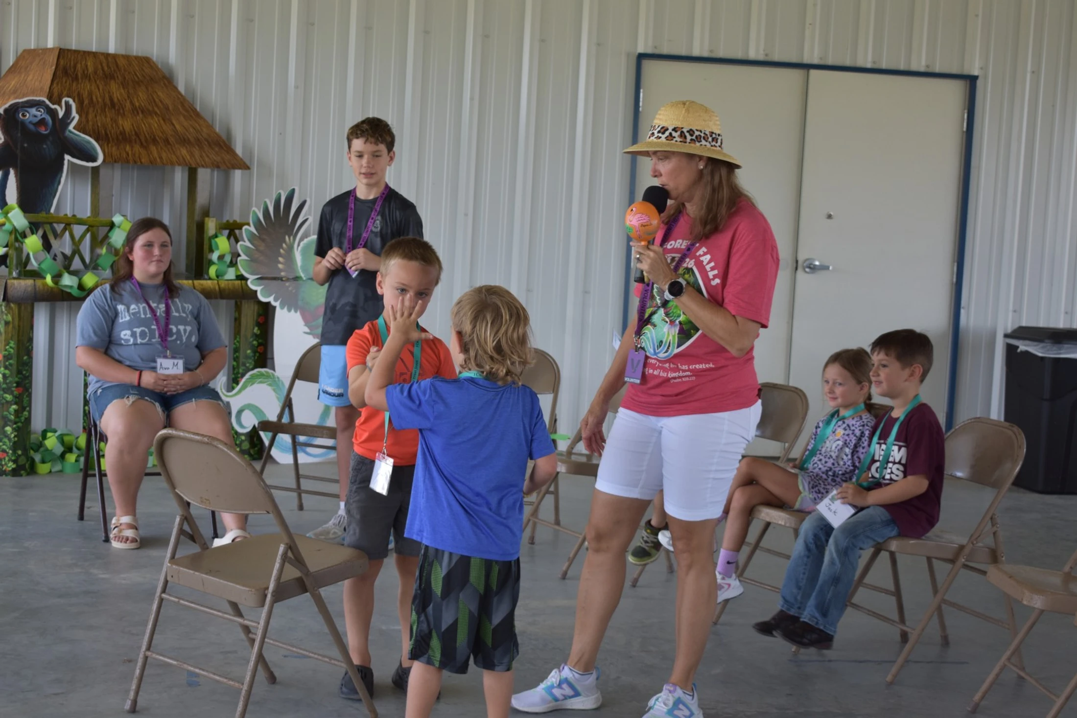 A woman in a straw hat holding a microphone talks to a group of children of various ages sitting and standing in a large room with jungle-themed decorations.