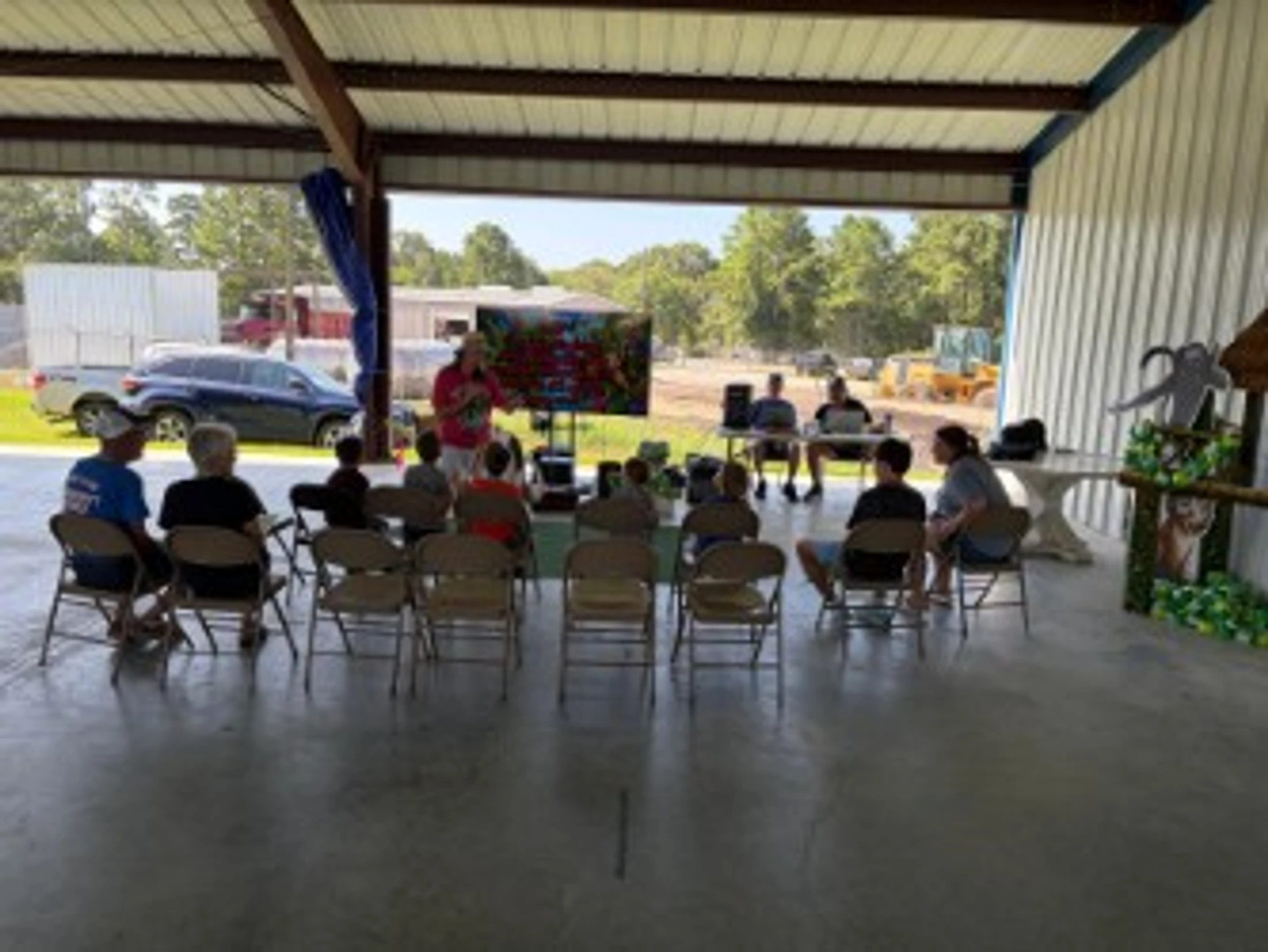 A group of adults and children sit on folding chairs in an open-sided building, watching a person give a presentation with a large screen.