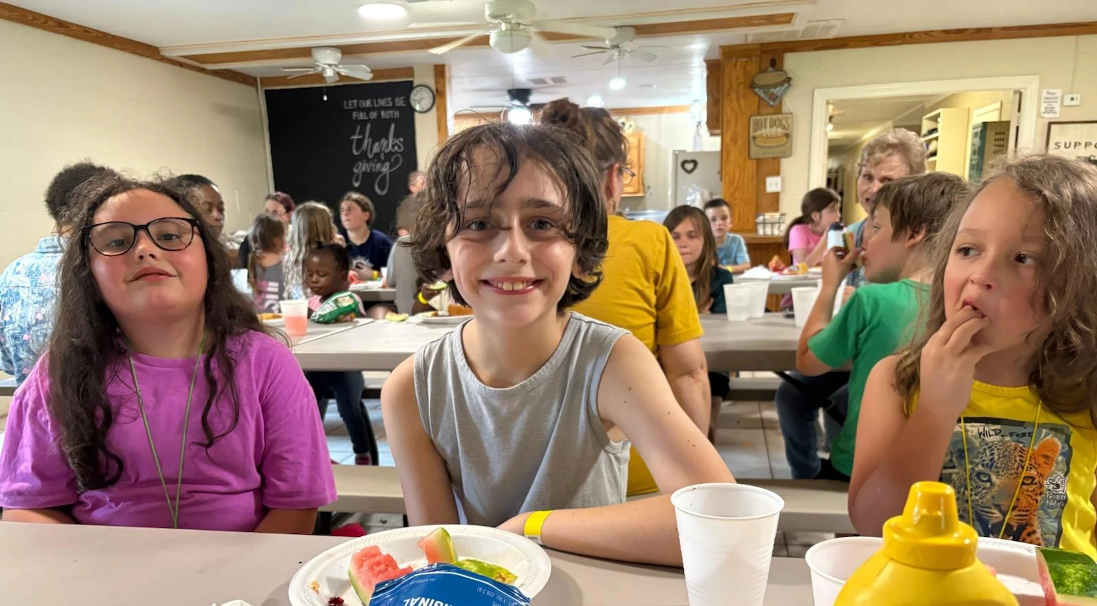 Three children sit side-by-side at a long table in a crowded cafeteria. The child in the middle smiles at the camera while the others eat their food.