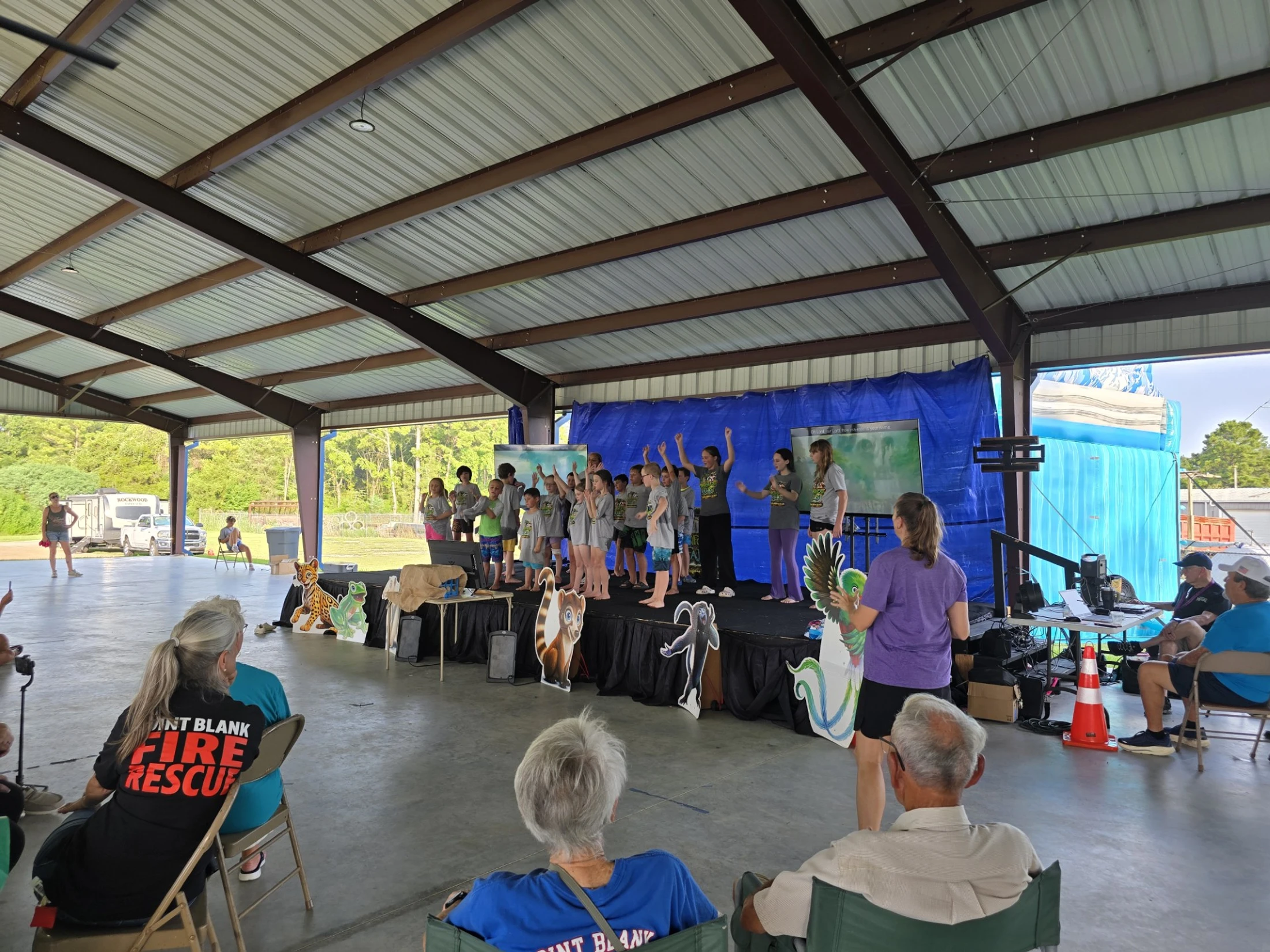A group of children in matching t-shirts perform on a stage for an audience under a covered outdoor pavilion, with cartoon animal cutouts in front of them.