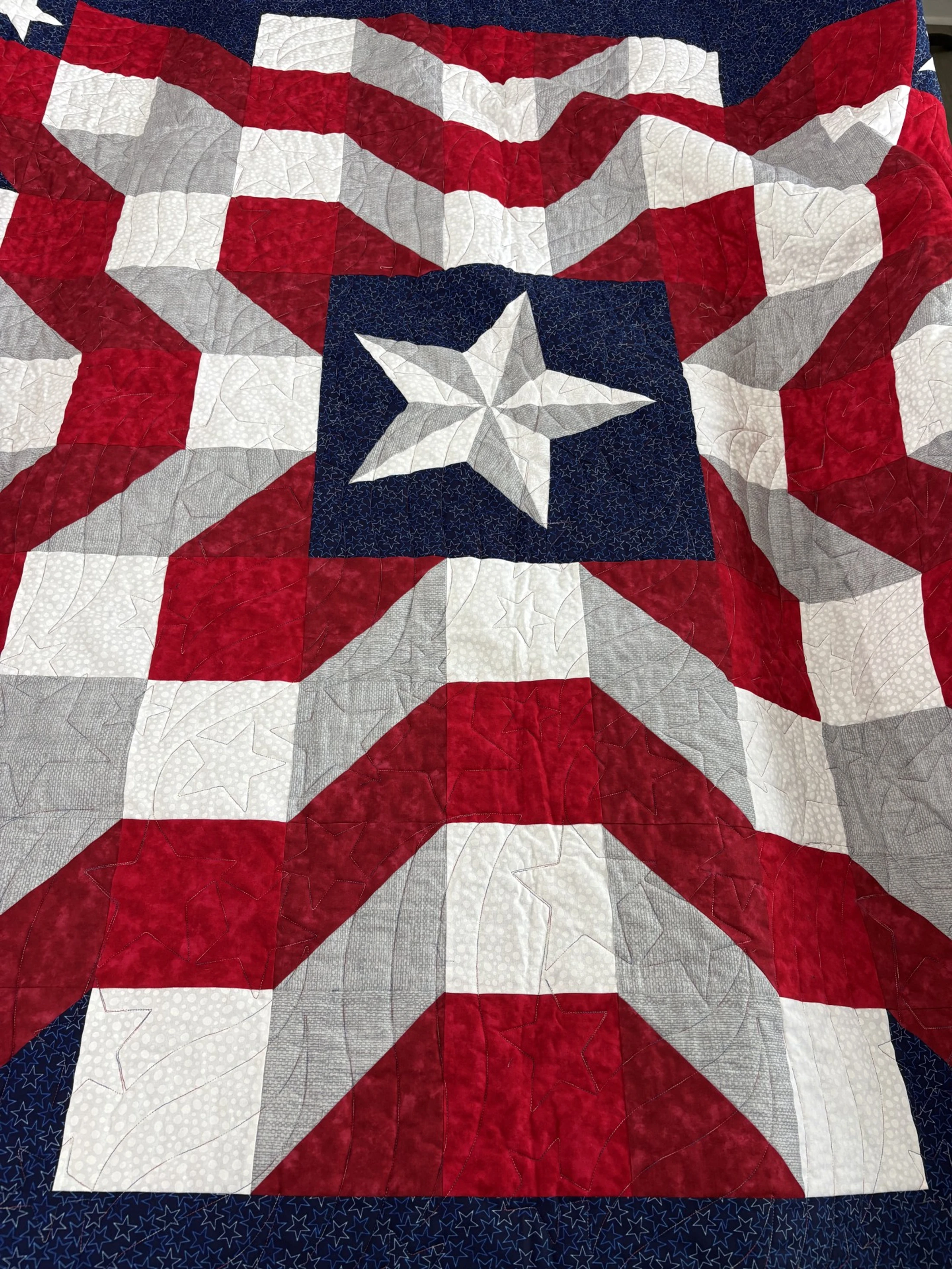 A handmade quilt with a patriotic theme. Red, white, and grey stripes radiate from a central dark blue square that contains a large, pieced star.