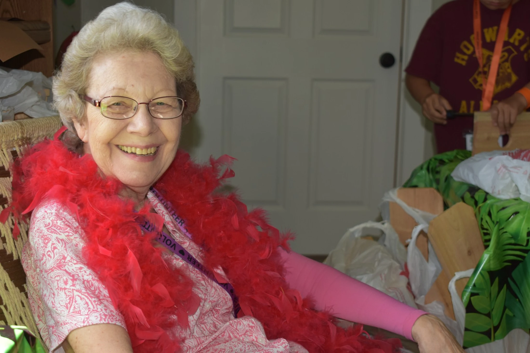 An elderly woman with glasses and short blonde hair smiles while seated. She wears a festive red feather boa over a pink patterned shirt.