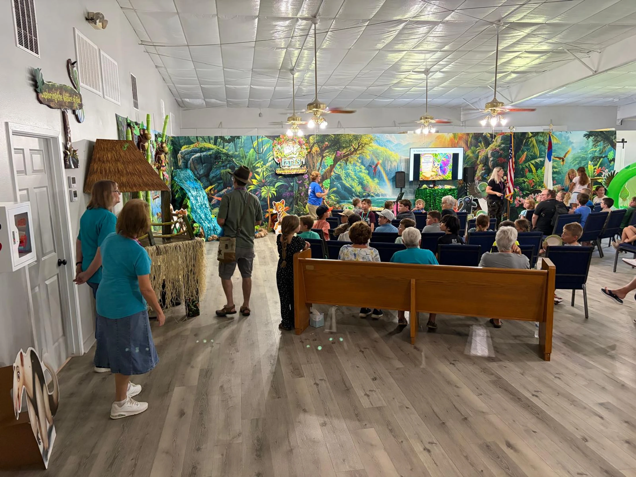 A large room decorated with a colorful jungle theme, filled with children and adults sitting in pews and listening to a presentation.