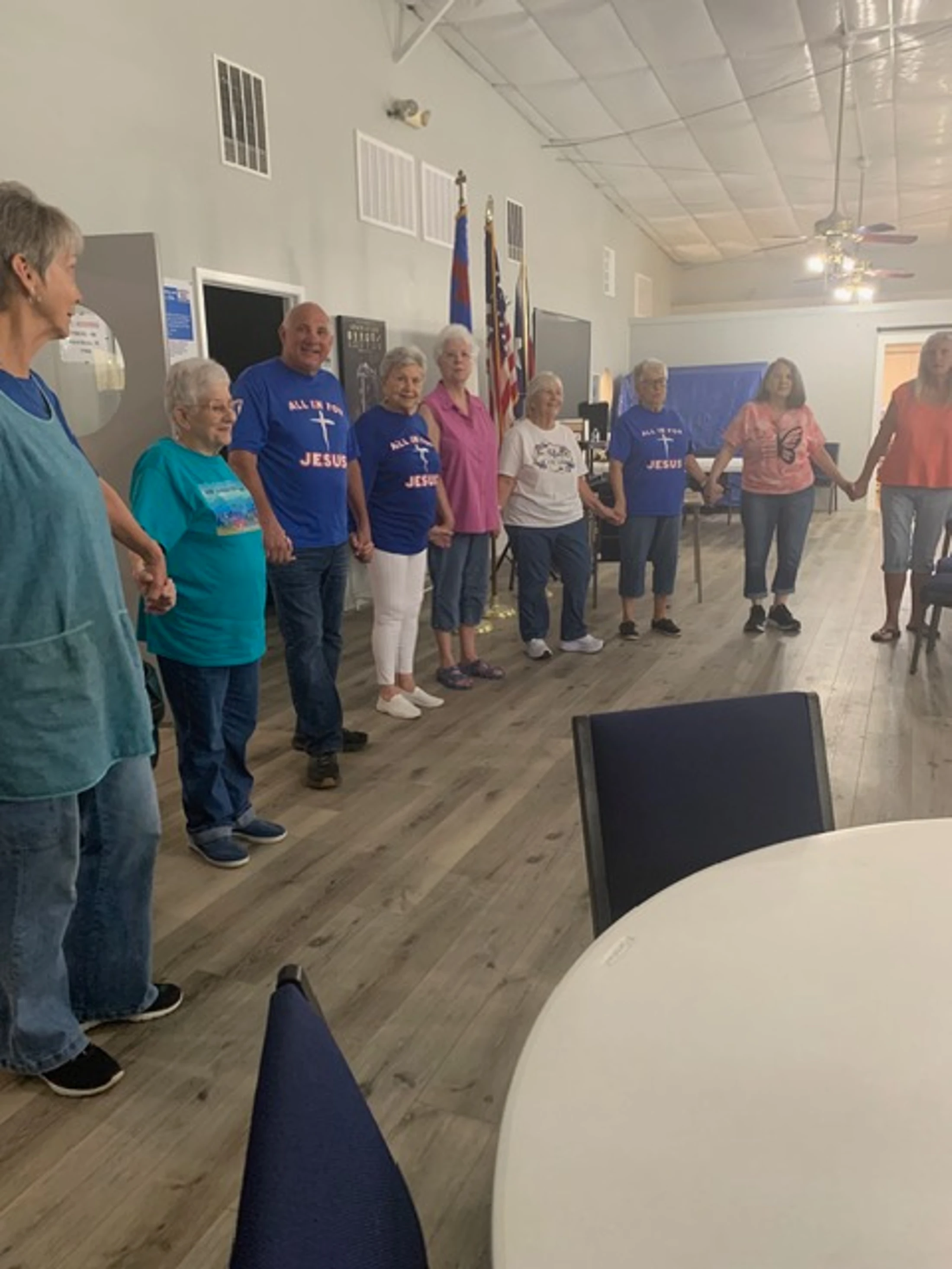 A group of older adults stand in a circle holding hands in a community hall. Several people wear matching blue t-shirts that say 'ALL IN FOR JESUS'.
