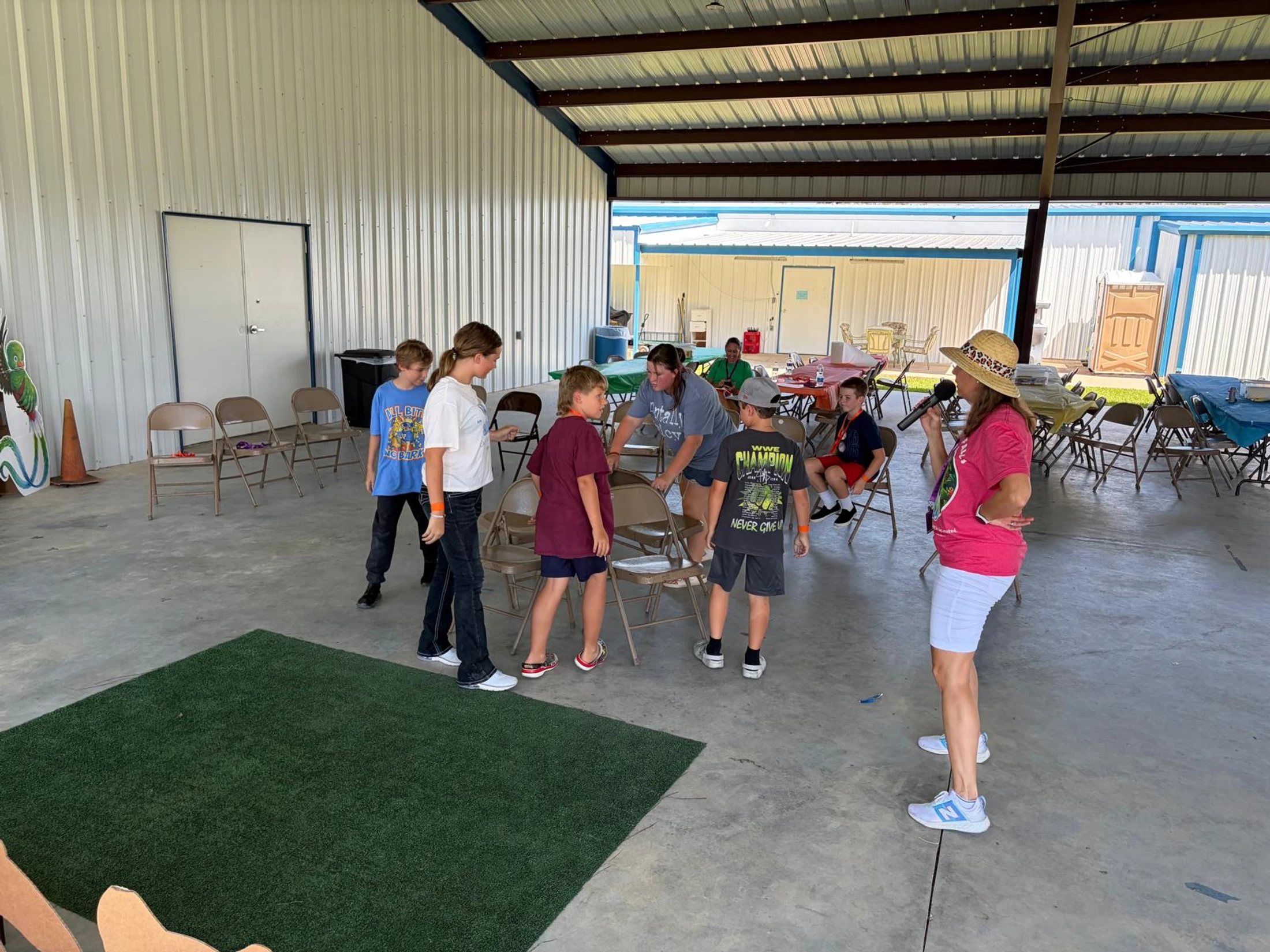 A group of children and adults stand around several folding chairs in a pavilion, preparing to play a game led by a woman holding a microphone.