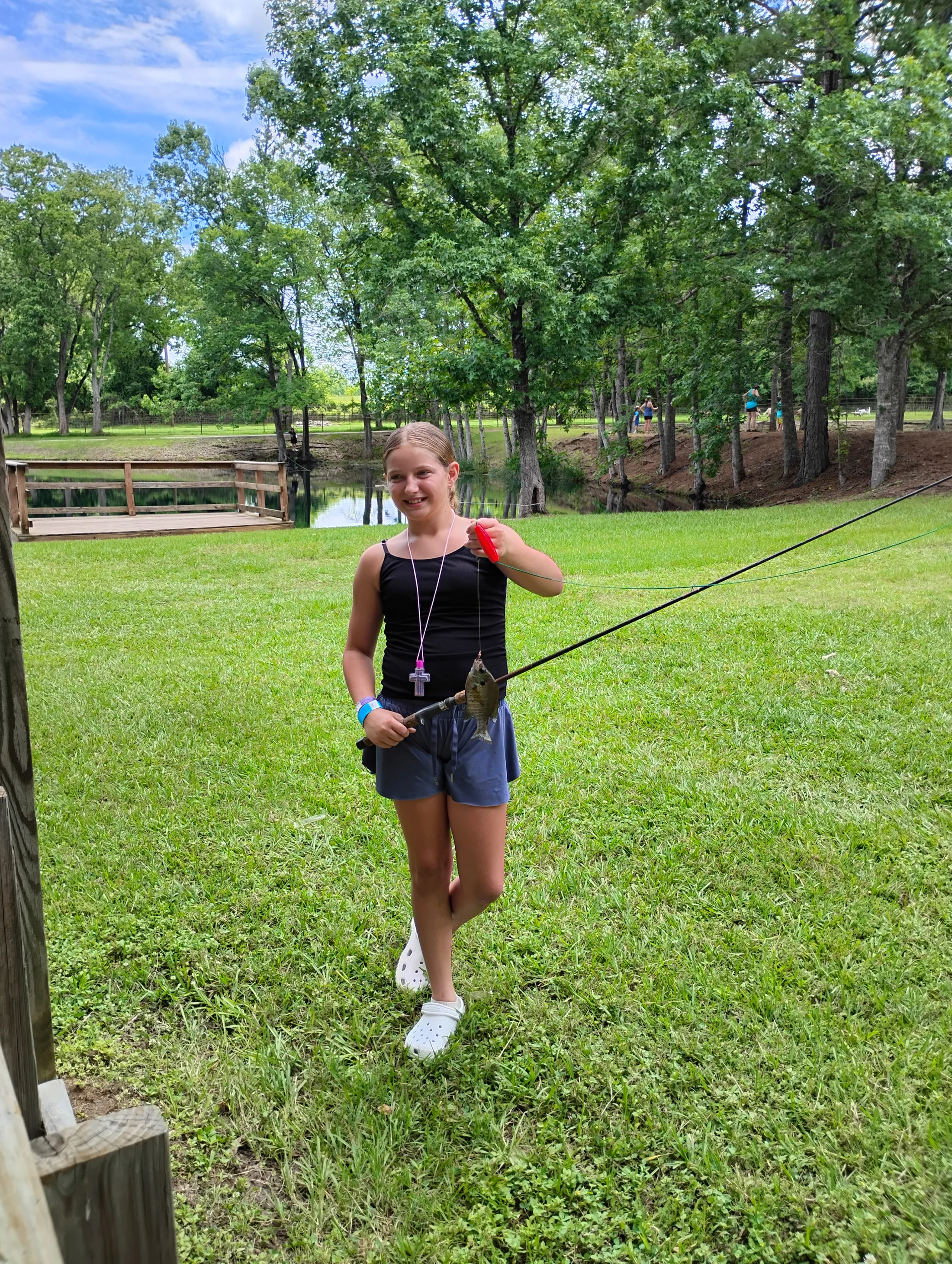 A smiling girl in a black tank top and shorts stands on a grassy lawn holding a fishing rod with a small fish dangling from the line.