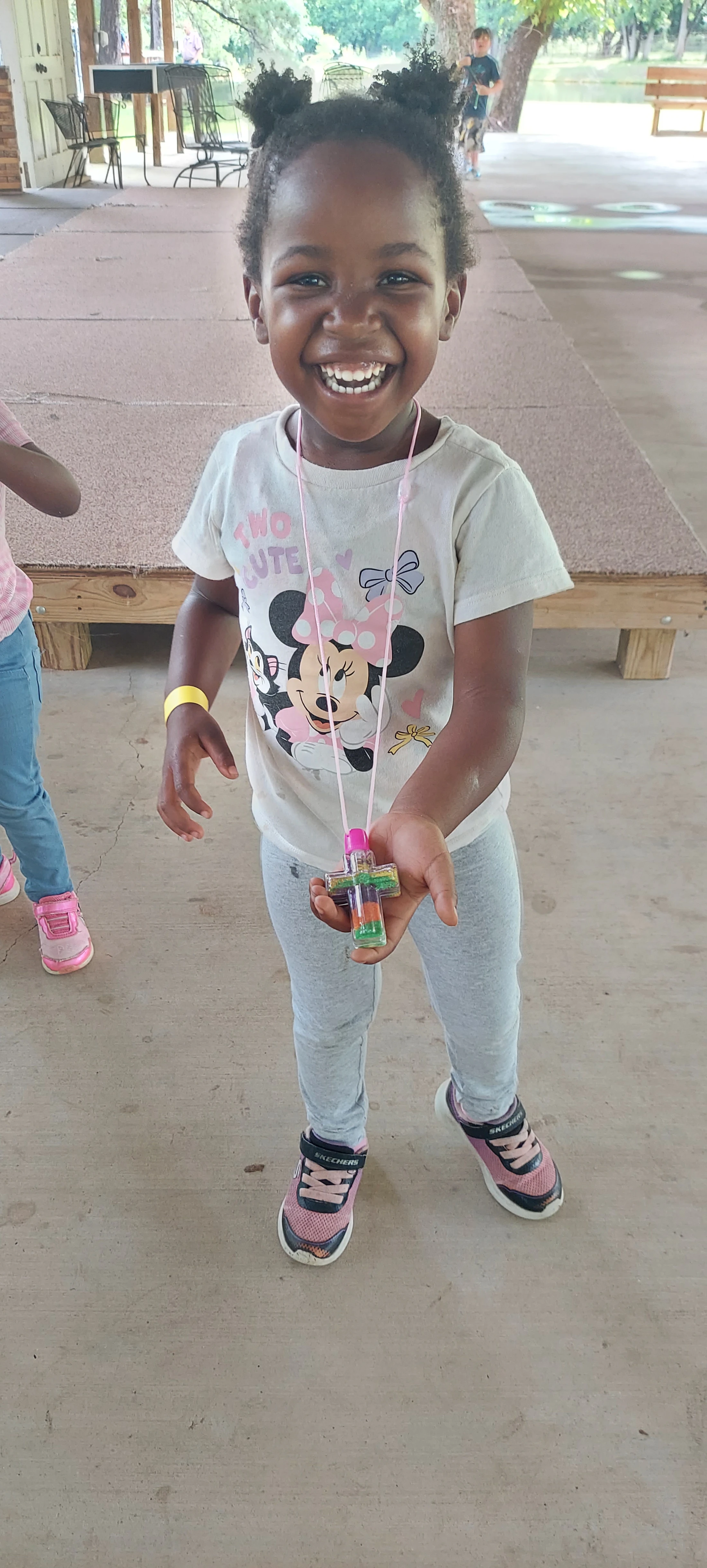A young Black girl with a big, happy smile holds up a colorful sand art cross necklace she is wearing. She's in a Minnie Mouse t-shirt outdoors.