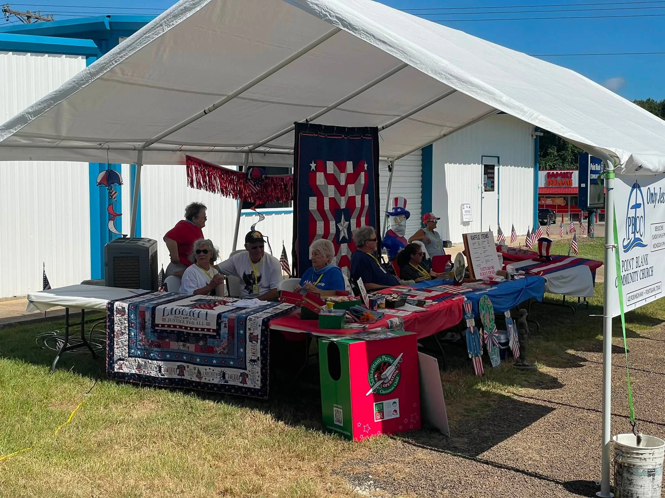 People gather at a booth under a white tent decorated with American flags and quilts for a Point Blank Community Church event on a sunny day.