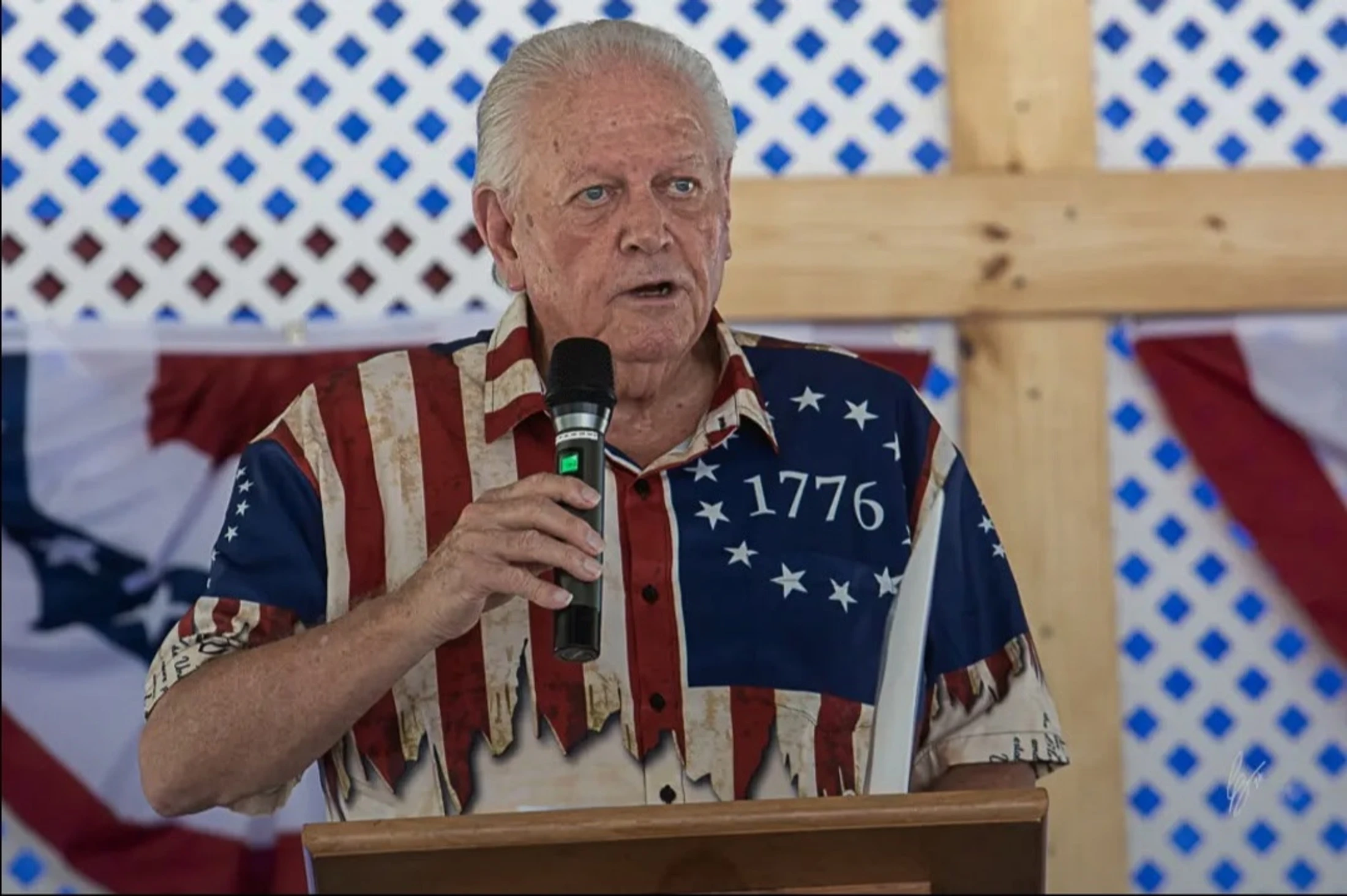 An older white man with gray hair speaks into a microphone. He wears a tattered American flag shirt with '1776' on it and stands at a wooden podium.
