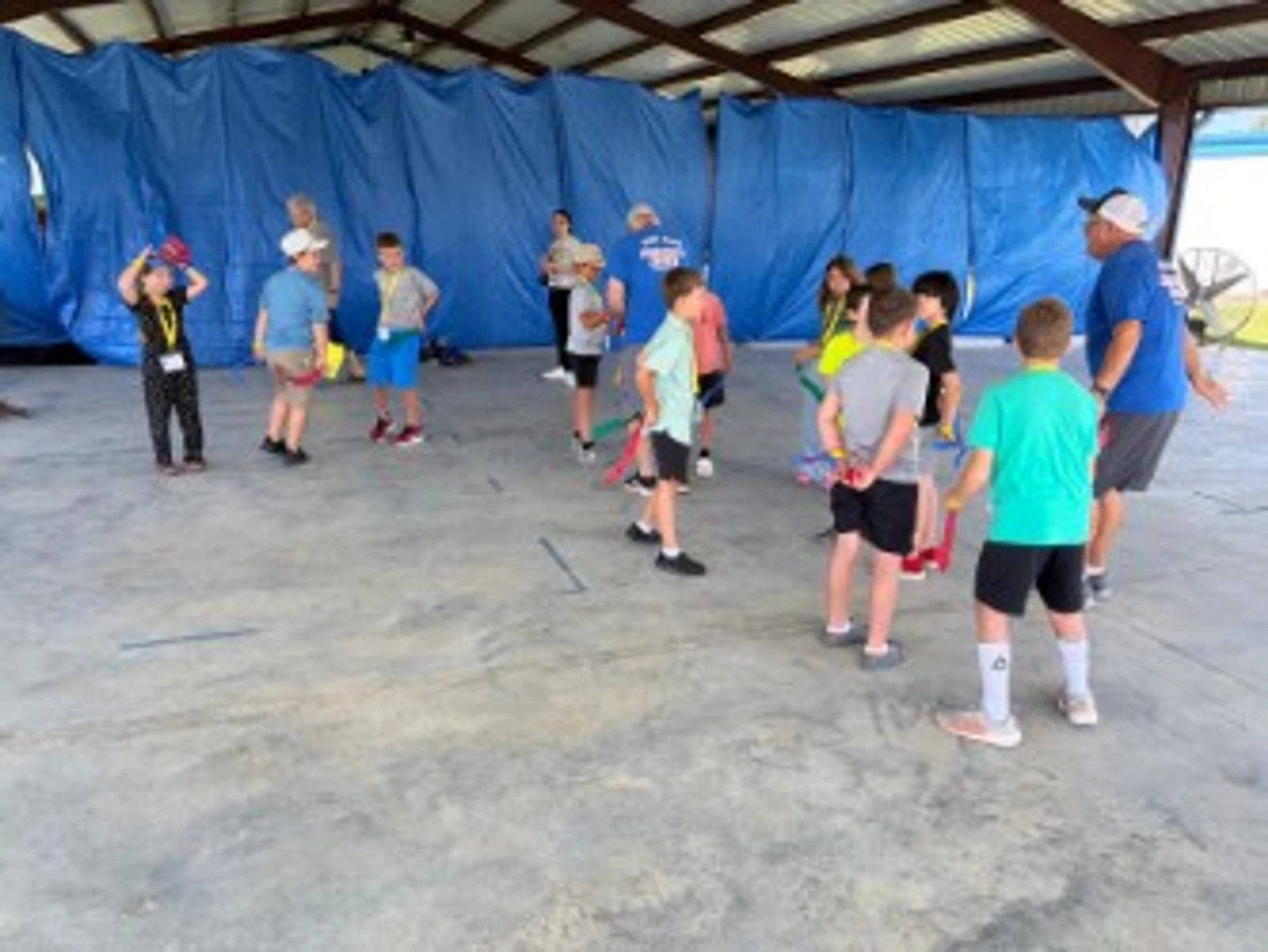 A group of young children and a few adults gather under a pavilion for an activity, holding colorful foam noodles while listening to an instructor.